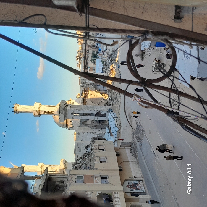 The image depicts a wide street scene in Khan Yunis, Palestinian Territory, under a clear blue sky with sparse clouds, illuminated by warm, low-angle sunlight. The environment is characterized by widespread structural damage.
In the center-background, a large mosque with two minarets and a central dome is prominently visible. The mosque is severely damaged, with large sections of its walls and structure collapsed, leaving piles of rubble at its base. Both minarets, though damaged, remain standing; small, dark figures are perched atop the right minaret.
To the left of the mosque, a multi-story building also shows extensive damage, including exposed interiors and missing sections. A mural featuring "Squid Games" and "Palin Games" is visible on its ground-level facade. A lone individual stands near this building on the street, and another person is visible on an upper floor, looking out from a damaged section.
The paved street in the foreground is largely clear of debris, but its edges are lined with rubble and fragmented construction materials. Multiple individuals are present on the street: a group of approximately three to four men walks away from the camera on the left side, another group of two is on the right side, and several other solitary figures or small groups are scattered further down the street, including one near a cart-like object on the right. A single person in red is visible further down the left side of the street.
The immediate foreground is partially obscured by out-of-focus elements, including tangled dark cables, power lines, and a rusty metal frame, indicating an elevated vantage point for the photograph, potentially from a damaged structure. The watermark "Galaxy A14" appears in the lower-left corner.