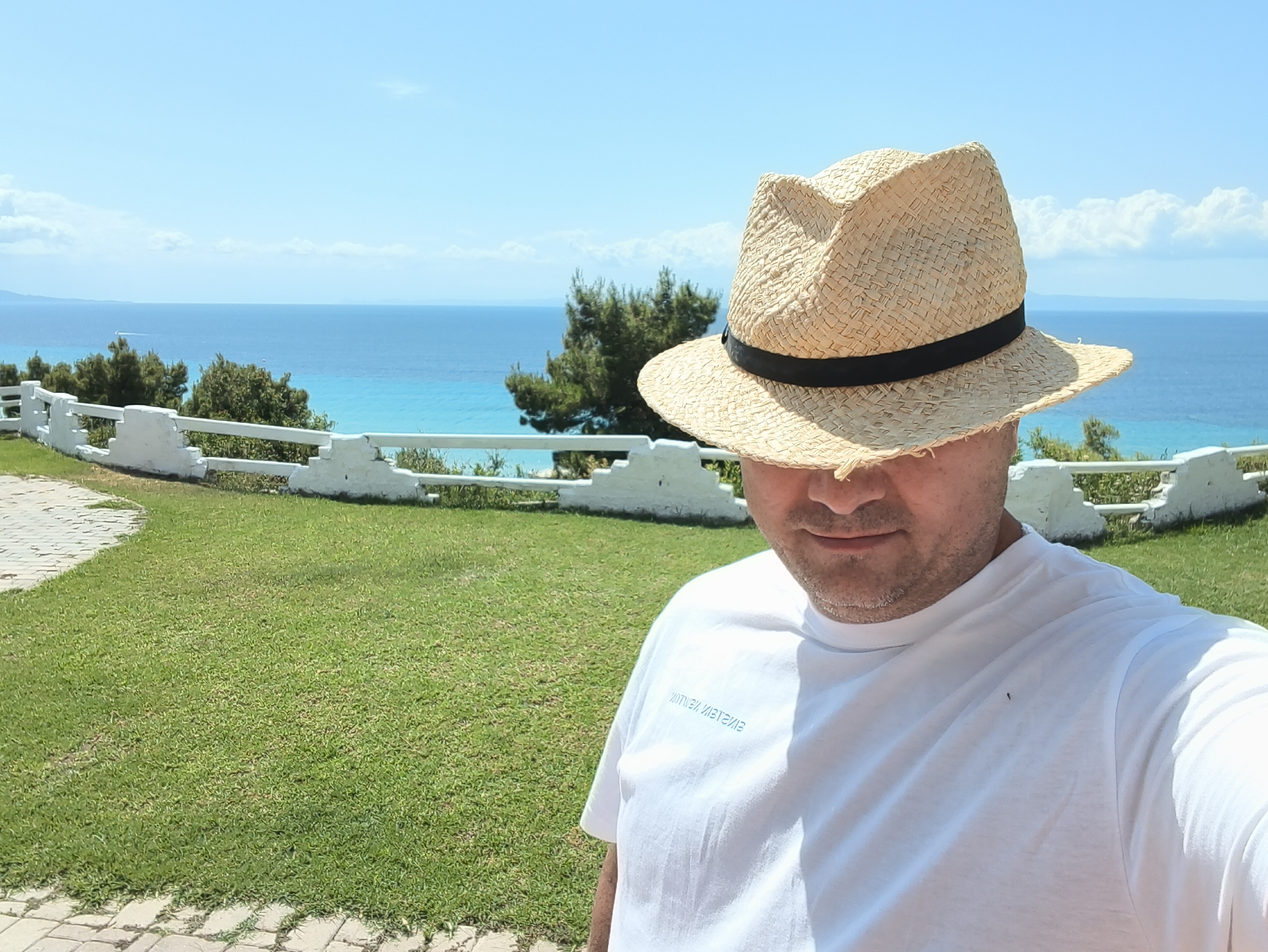 A man wearing a straw hat and a white t-shirt takes a selfie outdoors with a bright blue sky and the sea in the background. The man's face is mostly obscured by the hat, but his mouth is visible. He is standing on a grassy area, with a white railing and trees behind him. The sea is calm and clear, with a distant shoreline visible. The location appears to be a tourist area, possibly in Kassandra Municipal Unit, Greece, given the clear blue water and vegetation.