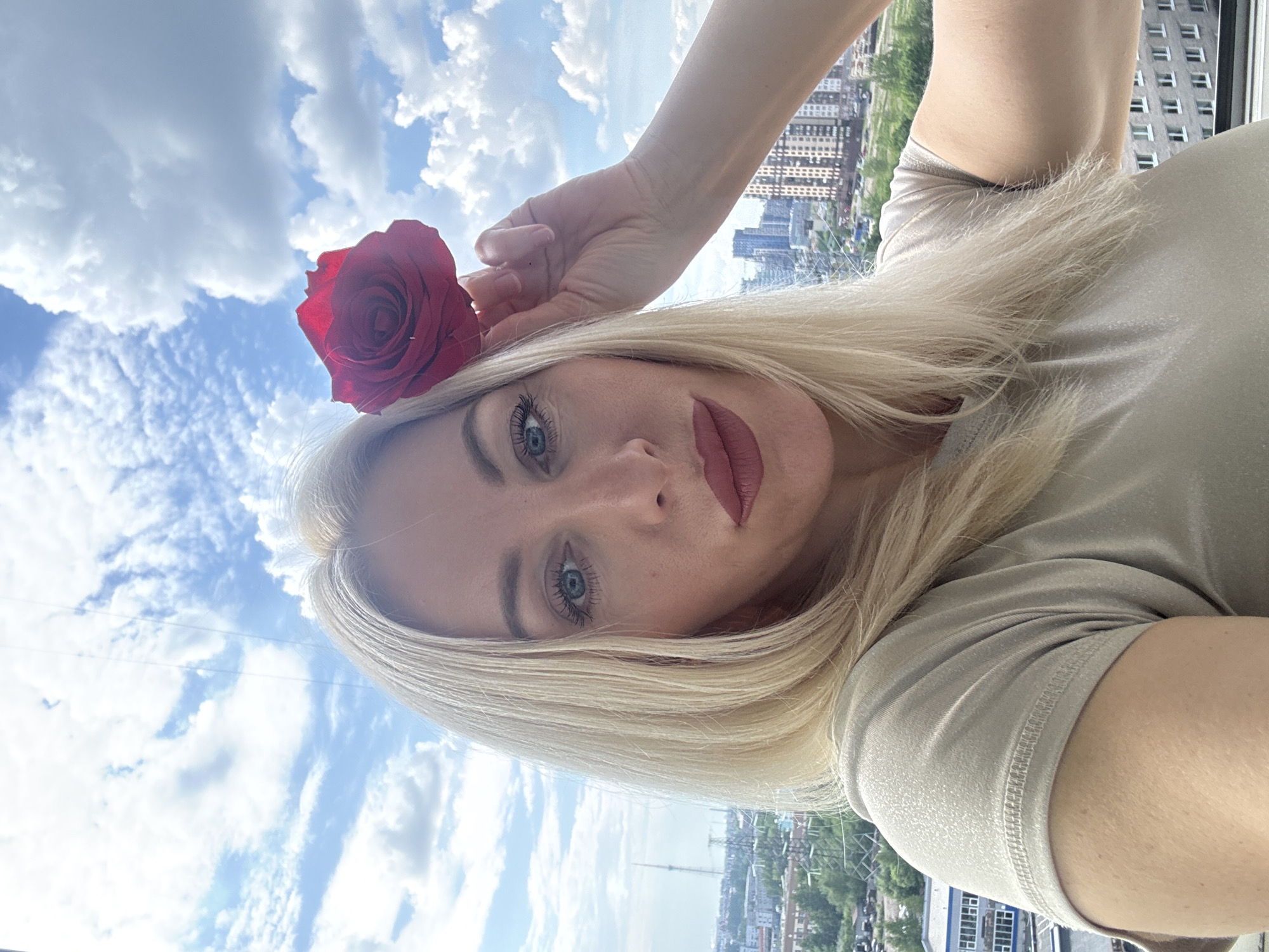 A woman is captured in a selfie-style portrait, looking directly at the viewer. She has long, straight blonde hair and prominent blue eyes, enhanced by dark mascara. She wears a reddish-pink matte lipstick. A vibrant red rose is tucked behind her left ear. She is wearing a light-colored, possibly beige or light grey, short-sleeved t-shirt, with her right arm and hand visible.
The background presents a bright urban landscape under a clear blue sky with scattered white, fluffy clouds. Below the sky, a panoramic view of a city is visible, featuring numerous multi-story residential buildings and some green trees. The perspective suggests the photograph was taken from a high vantage point, likely a balcony or window overlooking an urban environment. The time of day appears to be daytime, with sunny weather. There is no visible text in the image.