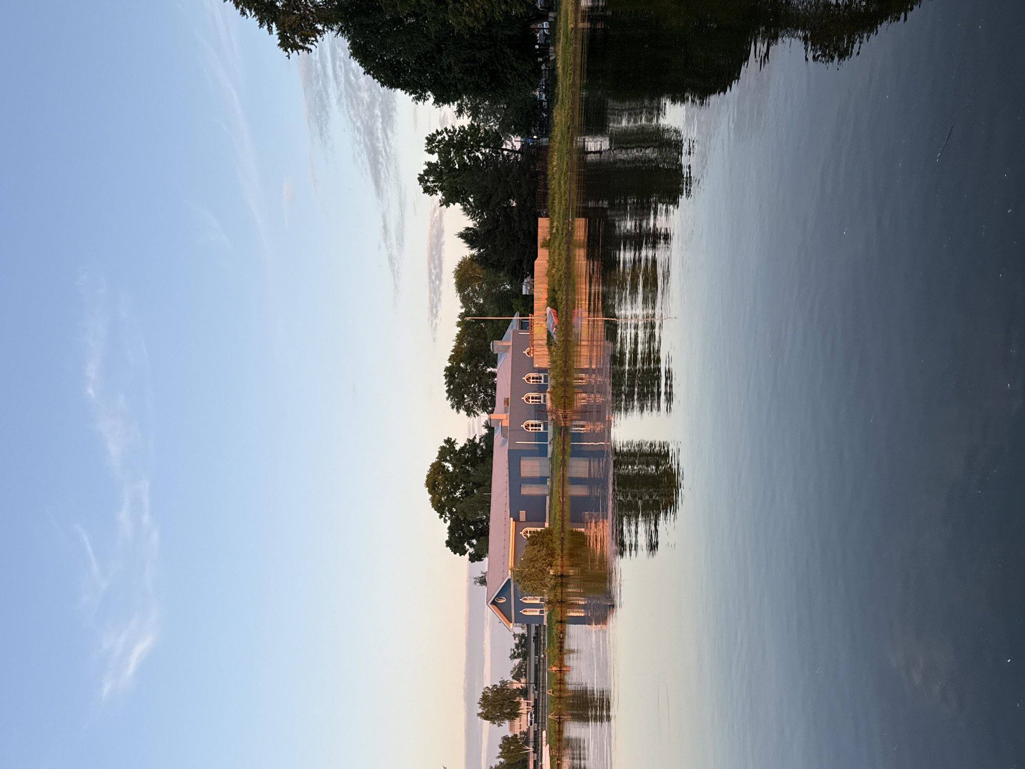 The image depicts a tranquil scene at what appears to be the Votkinsk Pond or a similar body of water in Votkinsk, Russia, during either sunrise or sunset. A long, light blue, single-story building with a light grey or white sloped roof is positioned on the far bank, its facade featuring multiple arched windows with white frames. A small triangular gable is visible on the left side of the building. The roof of the building and some elements on the shore reflect a distinct pinkish-orange hue, indicating the soft, warm light of the golden hour.
The water, occupying the lower half of the frame, is calm and dark, producing clear, mirror-like reflections of the building, the surrounding trees, and the sky. A narrow strip of green grass and low vegetation separates the building from the water's edge. To the right of the building, on the grassy bank, an overturned small boat, possibly with a red or orange hull, rests near a light-colored wooden fence. Various deciduous and coniferous trees, primarily dark green, are scattered behind and to the sides of the building, and along the right bank. In the far background, behind the building on the left, structures resembling a road or bridge are visible, along with the faint outline of vehicles.
The sky, occupying the upper portion of the image, transitions from a pale blue at the zenith to a subtle yellow and pink near the horizon, with sparse, wispy clouds scattered across it, some catching the soft, warm light. No individuals are visible in the scene.