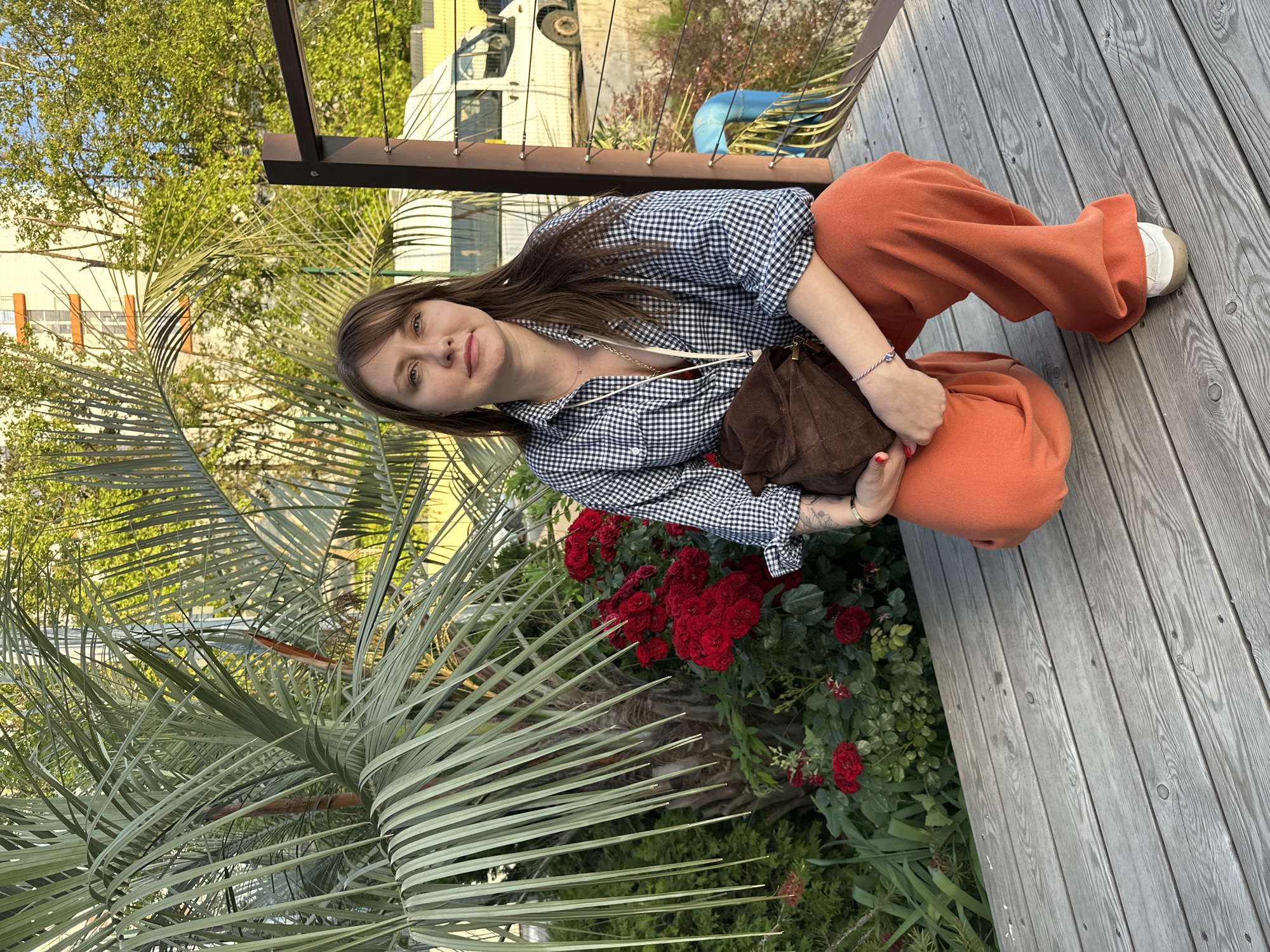 A young woman with long brown hair is squatting on a wooden deck in what appears to be a garden setting. She is wearing a black and white checkered shirt, loose-fitting orange pants, and white sneakers. A small brown purse is slung across her shoulder. She is looking towards the camera with a gentle smile.
In the background, there is lush greenery, including a large palm frond in the foreground, and several blooming red roses. Beyond the garden, a building and a white van are partially visible, suggesting an urban or semi-urban environment. The lighting suggests it is daytime, possibly late afternoon, given the warm tones. The scene appears relaxed and casual. There is no visible text in the image. The location context provided is Sochi, Russia.
