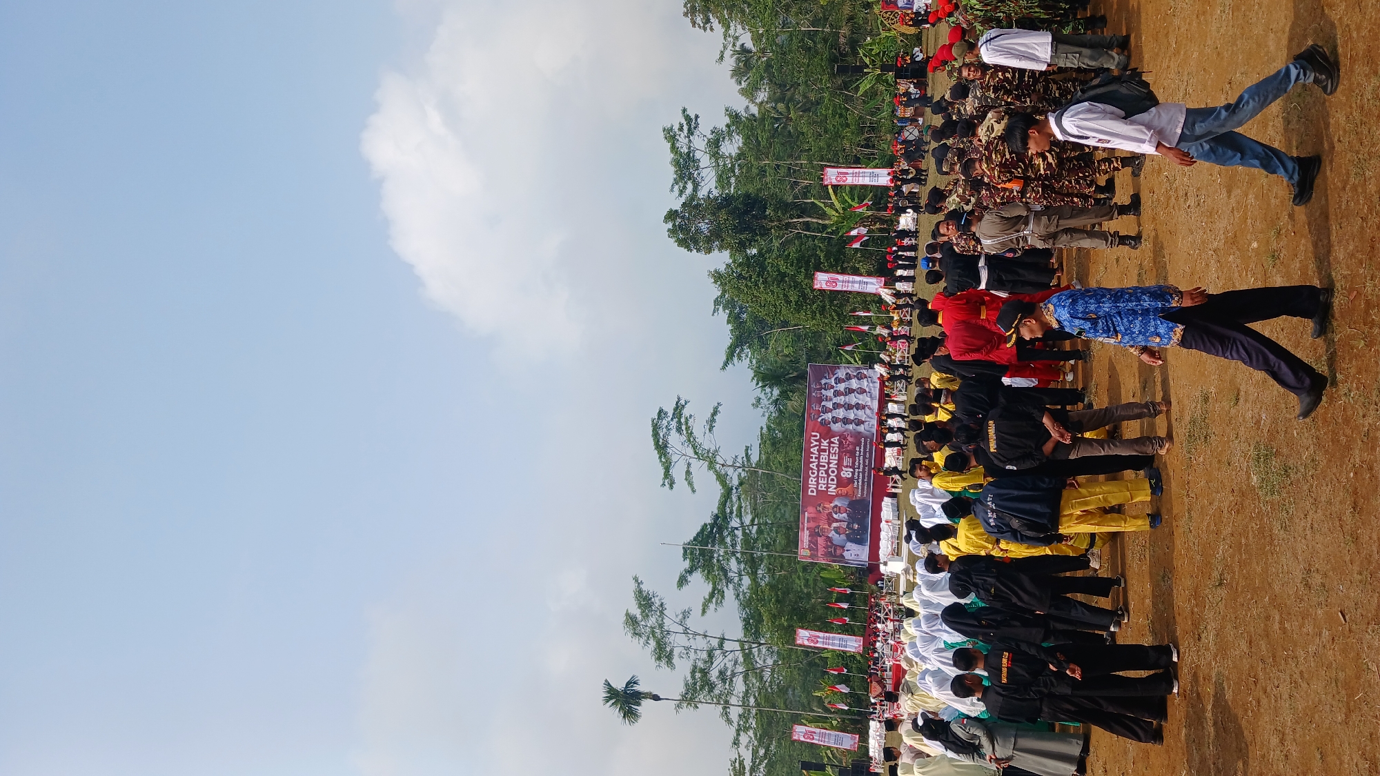 In Kepil, Indonesia, on a sunny day with a partly cloudy sky, an outdoor event is taking place. A large banner in the background reads "DIRGAHAYU REPUBLIK INDONESIA" and features images of people. A crowd of individuals, many wearing traditional Indonesian clothing or uniforms, are gathered on a dusty, brown field. Some are seated in rows of chairs, while others stand or walk around. The scene suggests a celebration or ceremony, possibly related to Indonesian Independence Day, given the banner's text. The environment is rural, with trees and vegetation visible behind the banner and people.