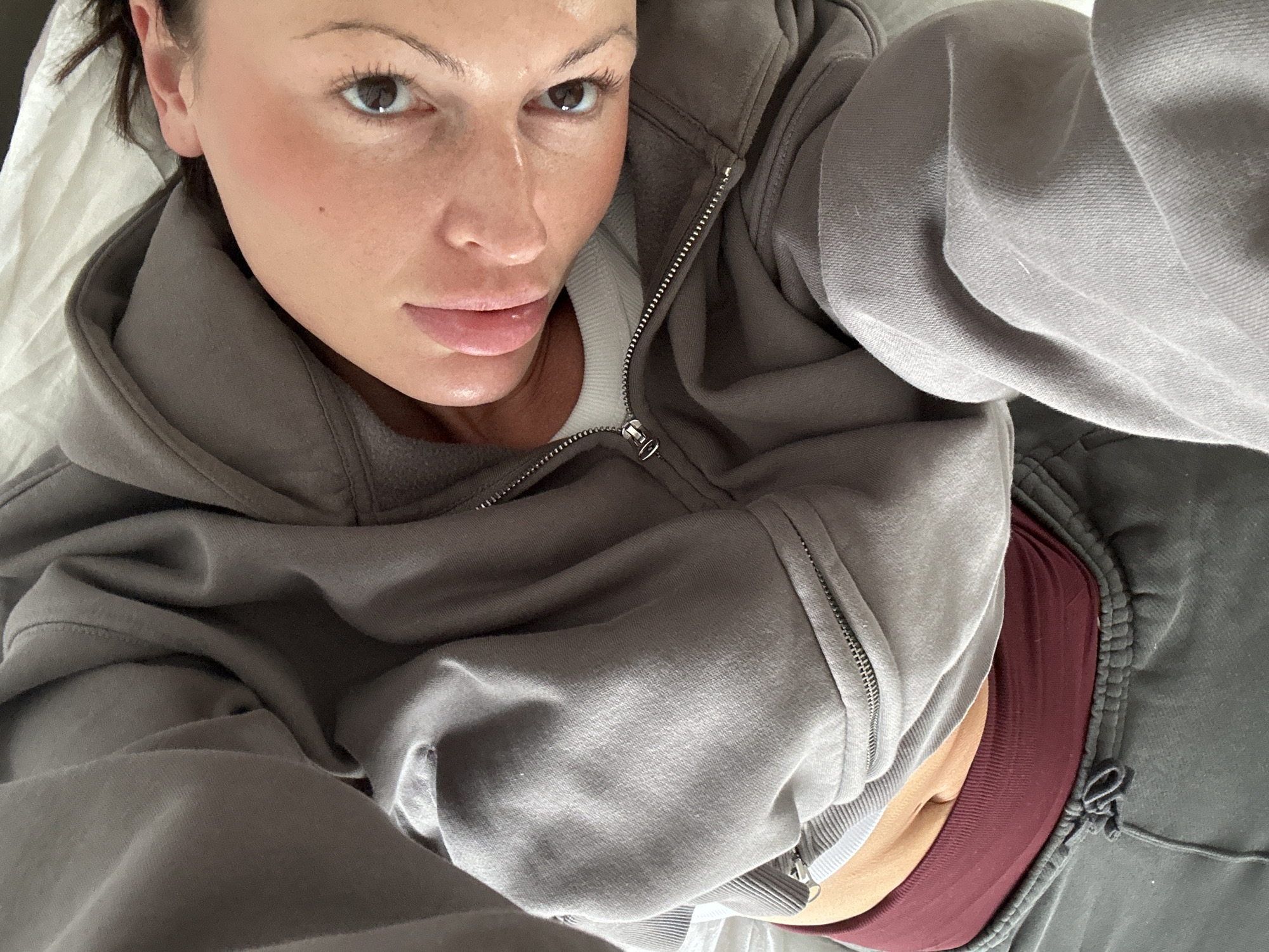 A close-up photograph depicts a female subject from an upward angle, likely taken as a selfie while the subject is lying down. The subject has fair skin, a natural flush on the cheeks, and a small dark mole on the upper right cheek. Dark eyes are open and gaze directly at the camera. The lips are full and slightly parted. Dark hair is visible near the right ear.
The subject is wearing a grey, full-zip hooded sweatshirt, partially unzipped to reveal a white crew-neck top underneath. Below the sweatshirt, a maroon or dark red elastic band, consistent with a sports bra or undergarment, is visible. The lower garment consists of dark grey sweatpants or joggers with a visible drawstring at the waist. The background behind the subject's head and upper body is a textured, wrinkled white fabric, consistent with bedding. The scene is indoors. The location is specified as Tunbridge Wells, United Kingdom, though no visual elements within the image confirm this geographical detail.