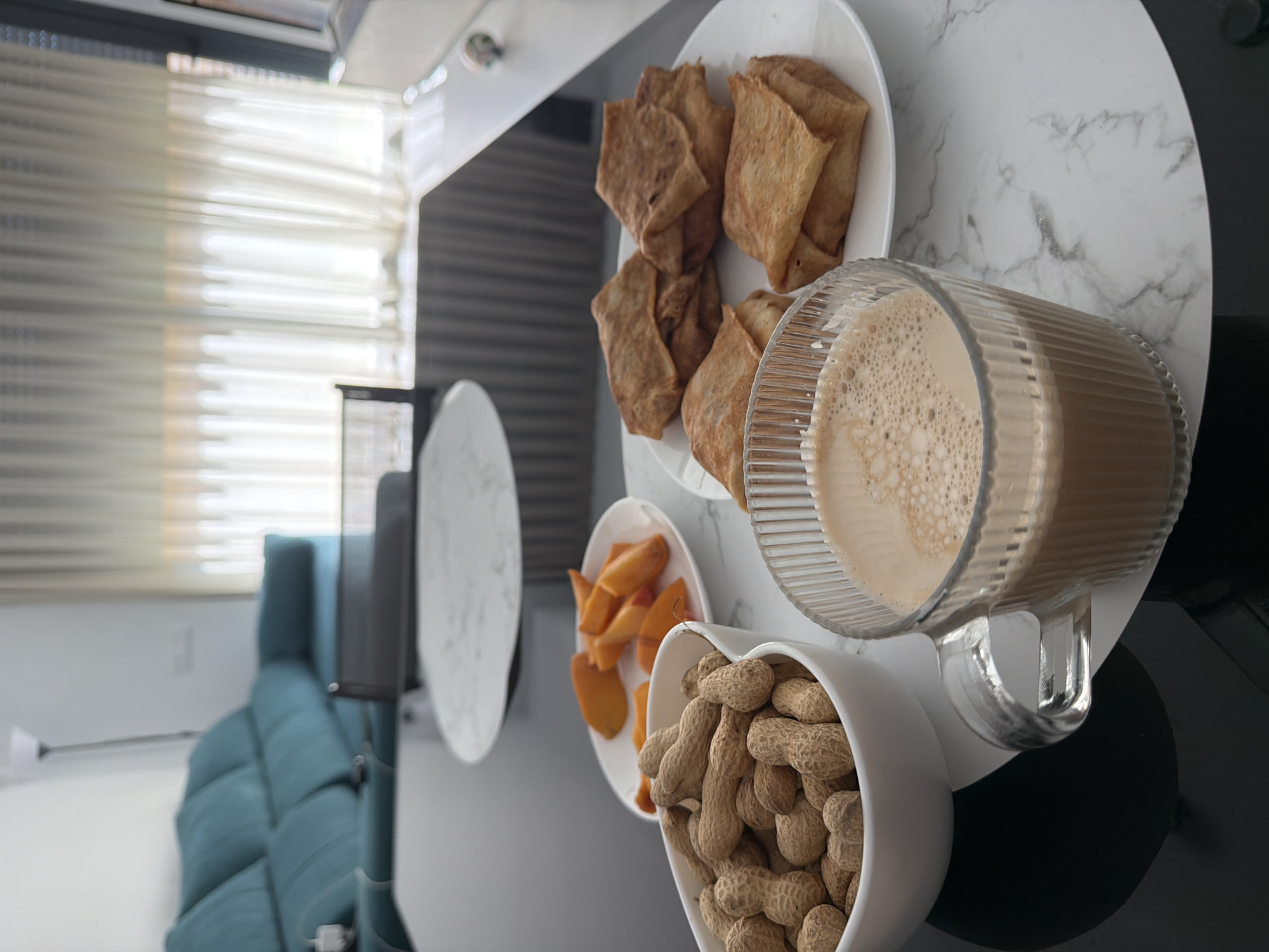 A close-up, high-angle shot captures a breakfast spread on a glossy black table, with a marbled placemat underneath. In the foreground, a clear glass mug filled with coffee and a frothy milk topping is positioned to the right. To the left of the mug is a white bowl overflowing with peanuts still in their shells. Behind the peanuts, a small white plate holds several slices of orange fruit. Further back, another white plate displays four folded crepes or pancakes. In the background, a contemporary living room is visible, featuring a turquoise sectional sofa and sheer curtains covering a large window. The setting is a modern apartment in Finestrat, Spain, likely a residential dwelling.