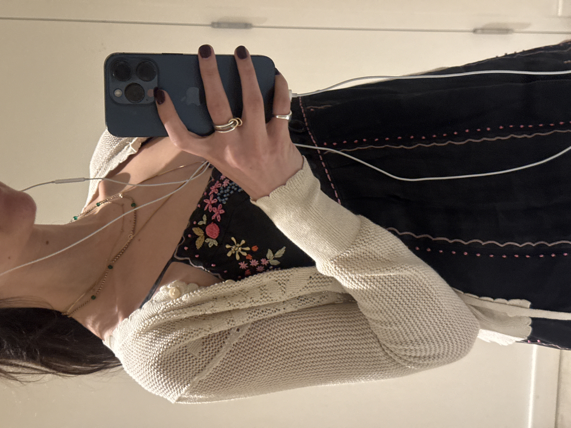 The image shows a person, only visible from the chest down, taking a selfie in a dimly lit room. The person is wearing a black dress with floral embroidery and a light-colored knitted cardigan. They are holding a dark blue smartphone in their right hand, which is adorned with two rings. The person's fingernails are painted a dark color. White earbuds are plugged into the phone and hang down the person's chest. The background appears to be a plain wall, possibly in a bathroom or bedroom, with a faint vertical line suggesting a door or a reflective surface. The lighting suggests artificial indoor lighting. There is no visible indication of the location within New York City, USA.