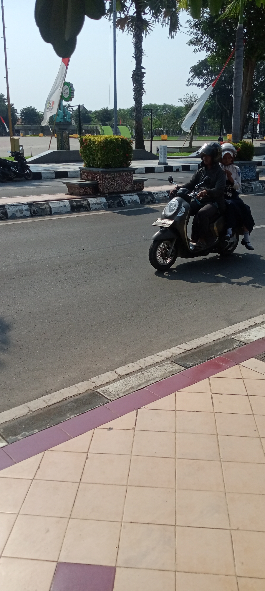 In Tuban, Indonesia, on a sunny day, a scooter carrying two people drives on the road. The driver, a man wearing a dark helmet and jacket, is focused on the road. Behind him, a young person, likely a student, wearing a white hijab and uniform, sits on the back. The scooter is a modern, dark-colored model with visible license plates. The road is paved, with a distinct curb separating it from a tiled pedestrian walkway. In the background, lush green trees and some foliage are visible, alongside what appears to be a public park or plaza with flags and decorative structures. The overall scene depicts a typical urban commute in Indonesia.