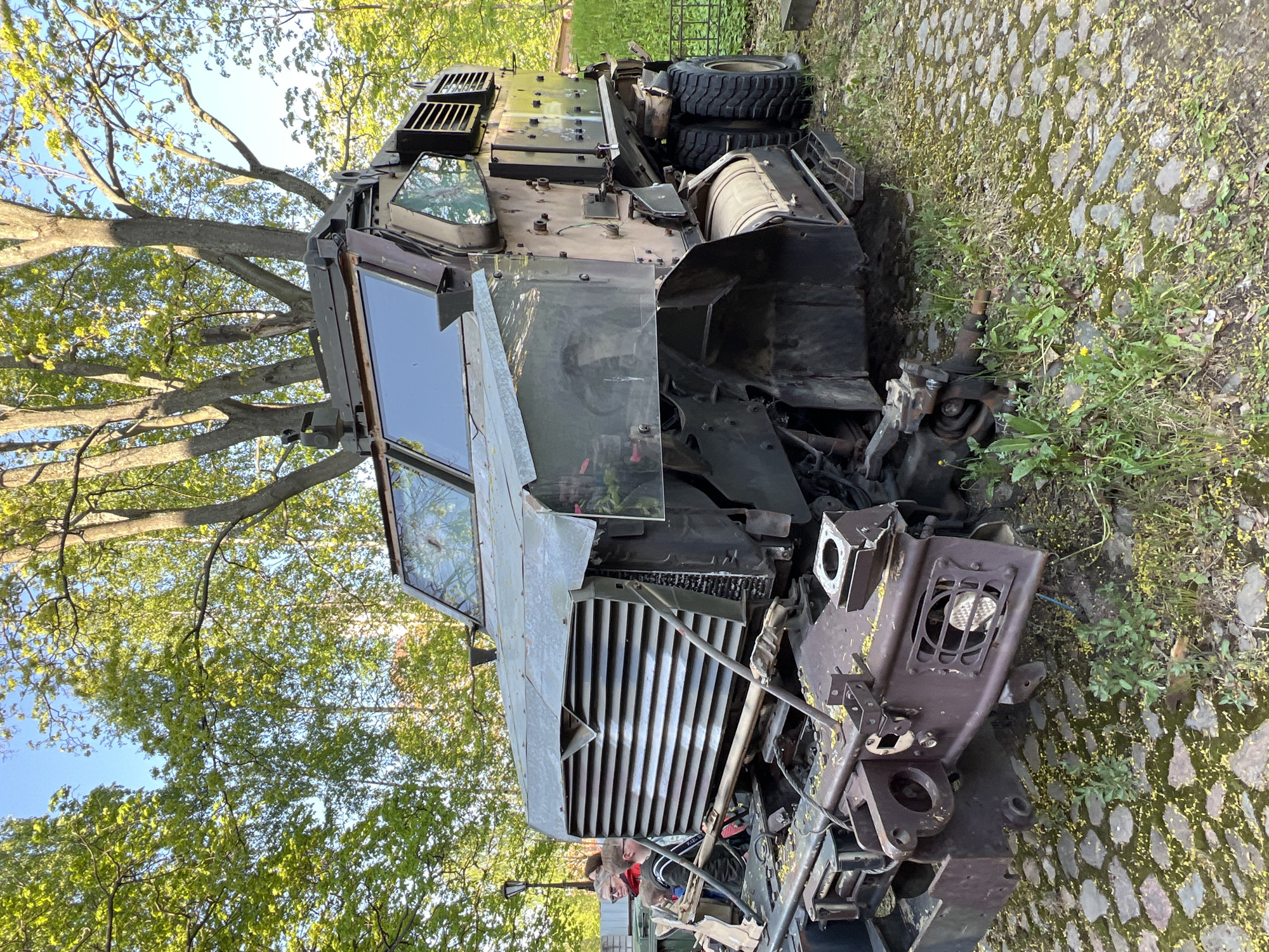 The image depicts a severely damaged, military-style vehicle situated on a cobblestone surface, possibly a street or plaza, overgrown with grass and weeds. The vehicle's front end is heavily crushed, with a mangled grill, shattered headlights, and severely distorted metal components exposing internal engine parts. The windshield and armored windows appear intact, though possibly scratched or damaged. The vehicle is painted in camouflage colors. A tree with green leaves is visible in the background and extends upwards and around the vehicle. Two people, wearing casual clothing and one with a red jacket, are partially visible inside the vehicle. There is no indication of the location being Saint Petersburg, Russia.