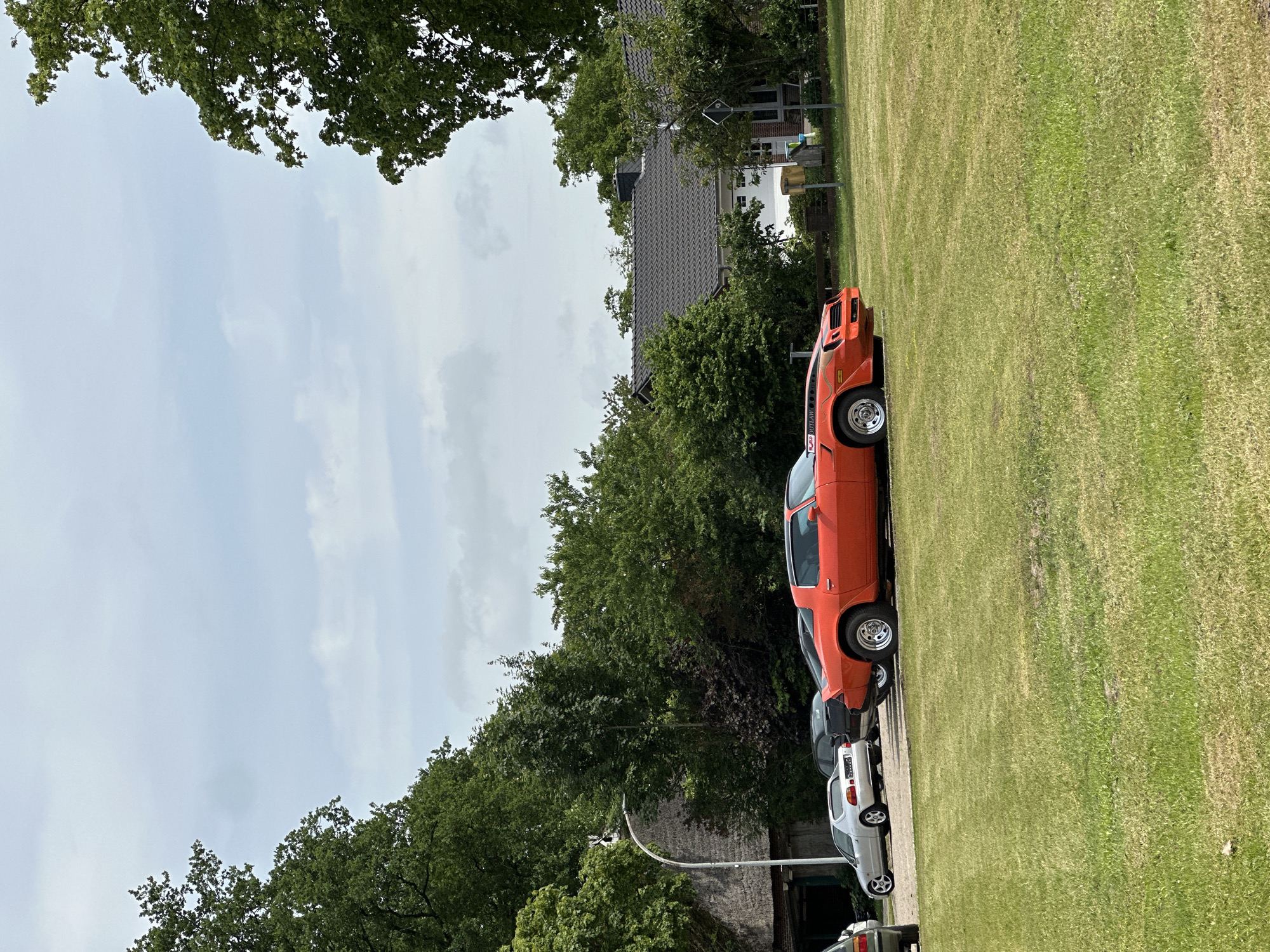 The image captures an outdoor scene in Kutenholz, Germany, under a cloudy sky. In the foreground, a vibrant orange 1980s Chevrolet Camaro, identified by "OUTLAW" branding on its side, is parked on a well-maintained green lawn. The Camaro has a distinctive black stripe running along its hood and a spoiler on the rear. To the left of the Camaro, a silver Honda CR-X is parked, and behind it, a dark-colored vehicle is partially visible.
Further into the background, a large building with a thatched roof and a modern house with a dark tiled roof are situated amidst lush green trees and bushes. A street lamp with a curved pole stands near the thatched-roof building. The environment appears to be a residential or semi-rural area, with a paved area behind the parked cars, suggesting a parking lot or driveway. There are no people visible in the image. The overall impression is a serene, daytime snapshot of vehicles and architecture in a German setting.