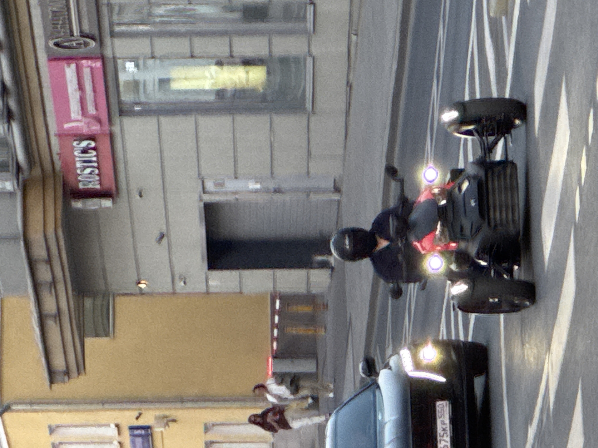 This image depicts a daytime urban street scene in Moscow Oblast, Russia. In the foreground and midground, a black three-wheeled vehicle with a red central section and illuminated headlights is seen moving across a white-striped crosswalk. The operator is wearing a black full-face helmet and a dark long-sleeved shirt. To the left of the three-wheeled vehicle, a dark-colored SUV or crossover is partially visible, displaying illuminated daytime running lights and headlights. Its front license plate, "675KP 550," is clearly legible, identifying its registration to the Moscow Oblast.
In the background, two primary buildings line the street. On the right, a multi-story building with a light grey horizontally banded facade features several windows and a ground-level entrance. A prominent red rectangular sign with white text reading "ROSTIC'S" is affixed to this building, accompanied by an adjacent pink sign and a round silver sign with a dark 'A' symbol. To the left, a multi-story yellow building with visible windows stands.
Two pedestrians are positioned on the sidewalk in front of the yellow building. The person on the left wears a dark top and light pants, holding an item in their hands. The person on the right wears a white top and light pants, carrying a shoulder bag. Behind the pedestrians, red and white striped barriers and two yellow bollards are visible, possibly delineating a restricted area. The road surface is asphalt, marked with white paint for the crosswalk.