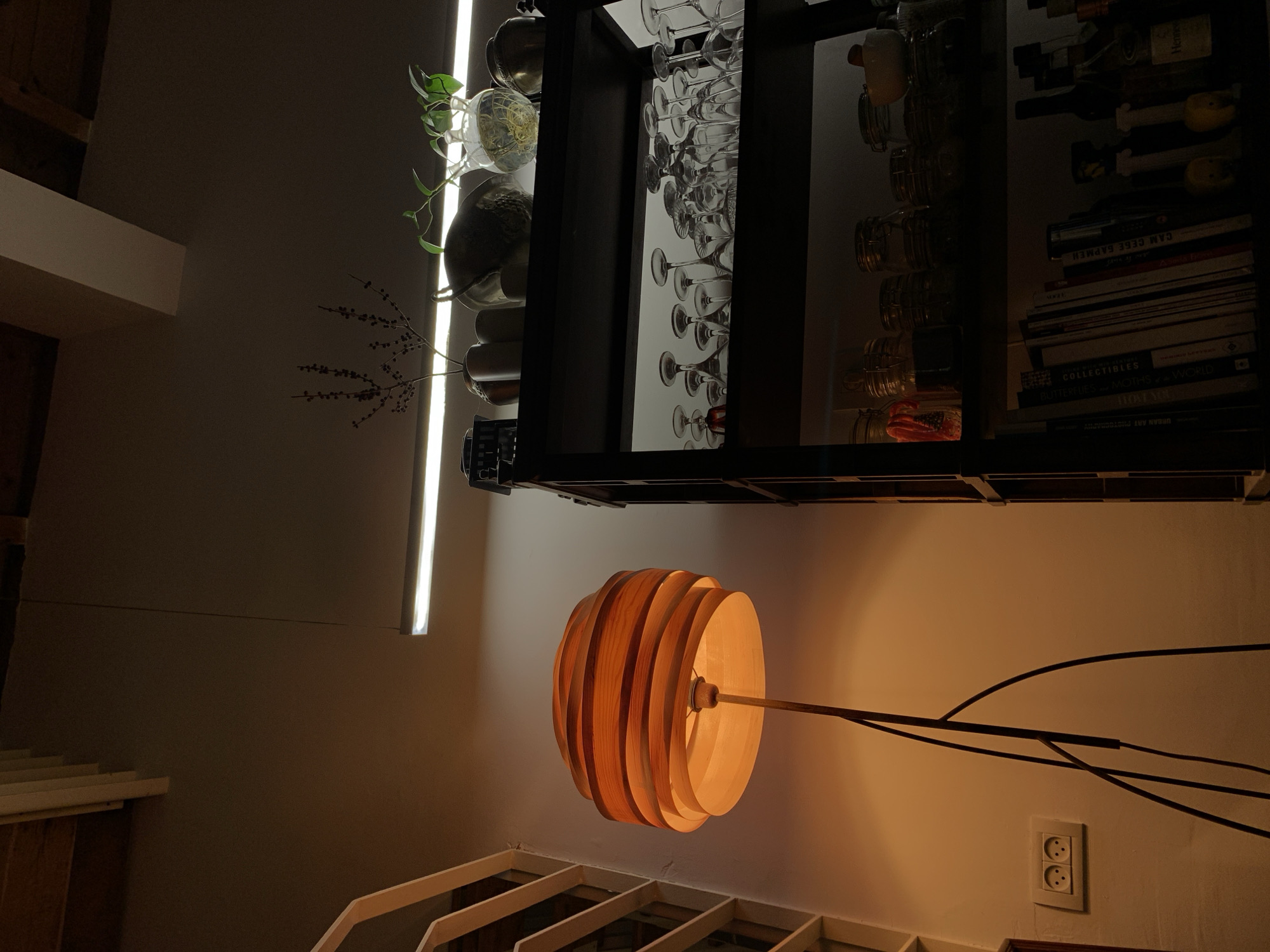 An indoor scene is captured from a low angle, depicting an interior space in Tel-Aviv, Israel, characterized by varied artificial lighting. On the left, a tall floor lamp with a multi-layered, circular shade constructed from horizontal wooden slats emits a warm, orange-toned light. Its dark, thin, branching metal stand is visible against a light-colored wall. Below the lamp's light source, a white electrical outlet with two circular sockets is mounted on the wall. Towards the upper left, exposed wooden beams or slatted ceiling elements are visible against a darker overhead surface. On the right side of the frame, a dark, open-backed shelving unit rises vertically. The top shelf displays several decorative items: various ceramic vessels, a metallic bowl, a clear glass vase containing a green plant with roots submerged in water, and a dried botanical arrangement in another vase. A prominent horizontal fluorescent strip light is affixed above this top shelf, providing bright white illumination. The second shelf down is populated with an array of inverted clear glass stemware, including wine glasses. The third shelf holds multiple clear glass jars with metal clamp lids. The lowest visible shelf contains a collection of books with diverse spine designs, alongside several bottles, one clearly labeled "Hennessy" among others. No people are present in the scene, and no explicit actions or interactions are occurring.