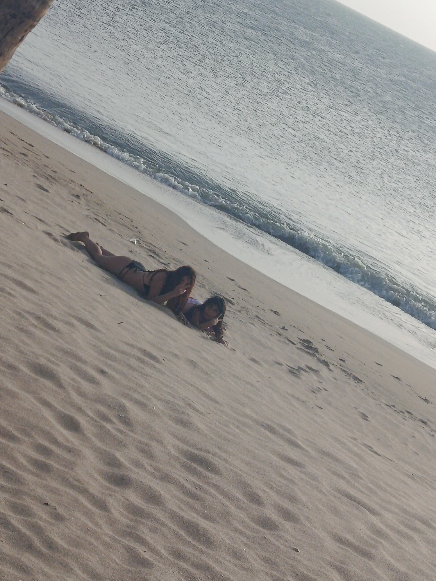 Two women are lying on a sandy beach. They are positioned near the shoreline with the ocean behind them. The location is Juan Griego, Venezuela.