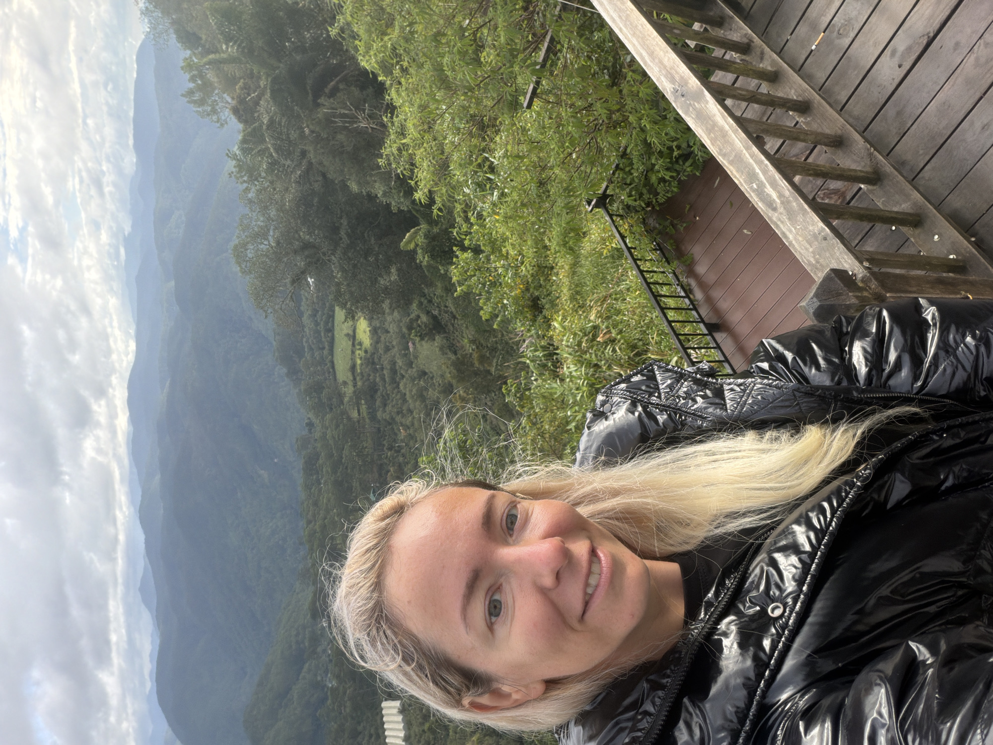 A young woman with blonde hair smiles at the camera, wearing a shiny black puffer jacket. She is standing on a wooden deck overlooking a valley filled with lush green trees and rolling hills. The sky is cloudy, with patches of blue visible. The scene appears to be a scenic overlook, possibly in a mountainous or hilly region. The woman is the primary subject, and the landscape provides the context. The image conveys a sense of outdoor exploration and enjoyment of nature. The location is not specifically identified as Phát Chi, Vietnam.