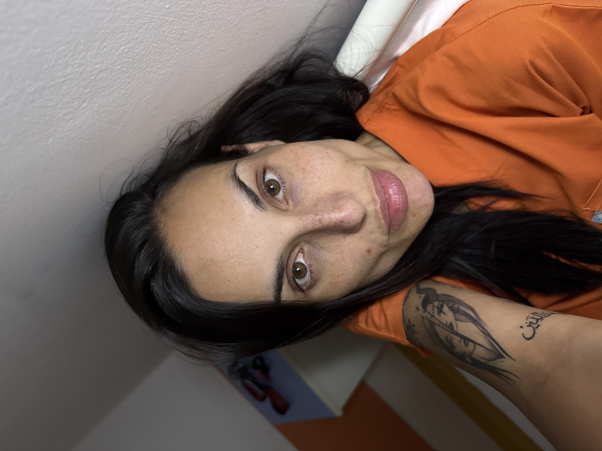 A woman with dark hair and brown eyes is lying down. She is wearing an orange top. She has a tattoo on her left arm, which appears to include Arabic script and the image of two faces. The background includes a white wall and part of what looks like a white bed or cushion. A partially visible bedside table is also present with some items on it. The location is not evident from the image, so the city of Pleven, Bulgaria, cannot be confirmed.