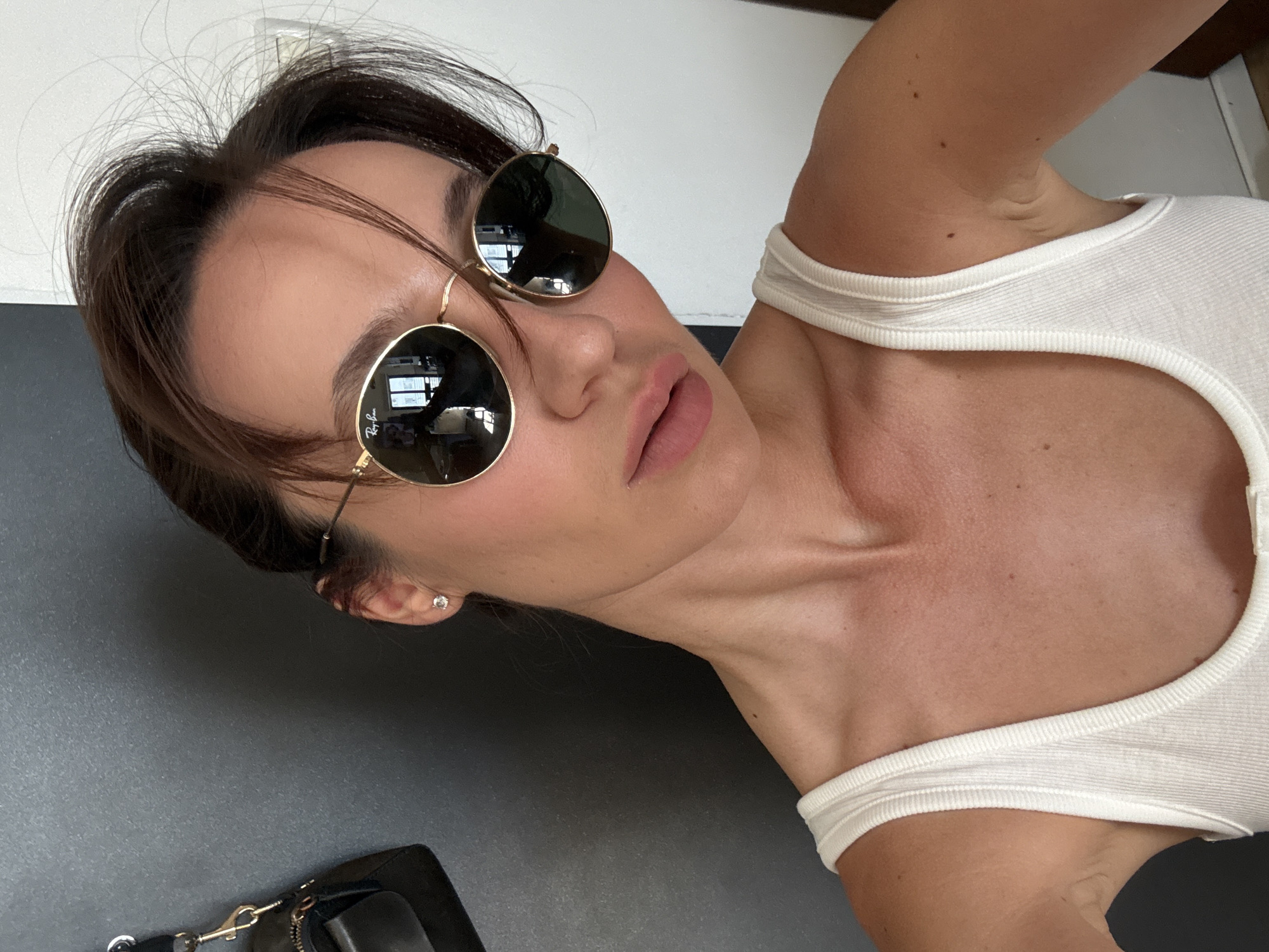 A woman is taking a selfie indoors, possibly in Tel Aviv, Israel. She is wearing a white tank top and round, gold-rimmed sunglasses. Her hair is dark and pulled back loosely. She has light makeup on, with blush on her cheeks and a light pink lipstick. A small diamond stud earring is visible in her left ear. In the background, a black bag hangs on a dark gray surface, and a white wall is visible to the right. The lighting suggests it is daytime.