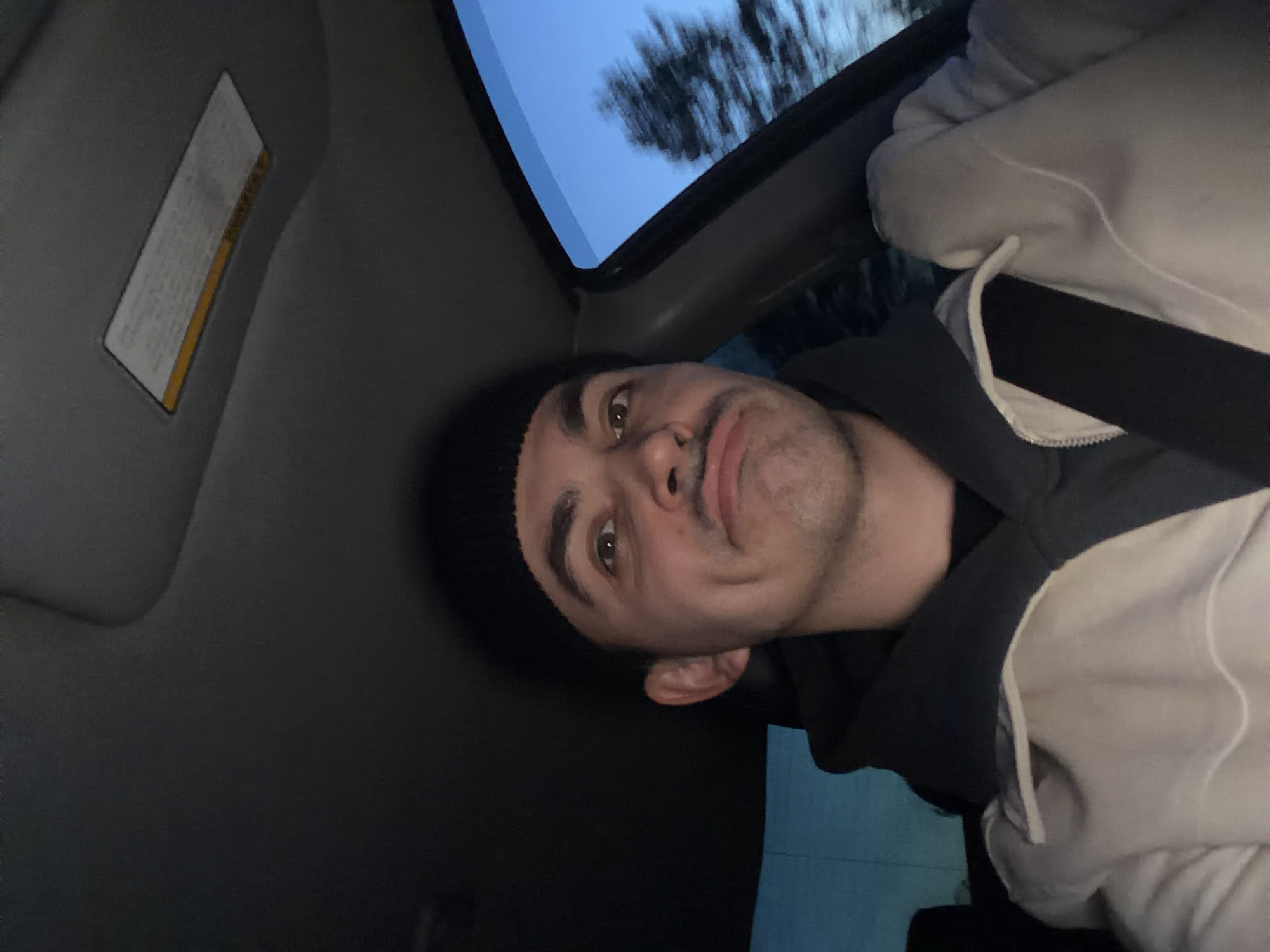 A selfie-style photograph, likely taken inside a vehicle. The subject, a young man with a black beanie, dark eyebrows, light skin, and stubble, is positioned in the driver's seat. He appears to be wearing a dark hooded sweatshirt and a white jacket/hoodie over it. He is wearing a seatbelt. The interior of the vehicle is visible, showing a sun visor, a partially visible sunroof with a view of trees and a blue sky. The location and city of Shaberdino, Russia cannot be determined from the image.