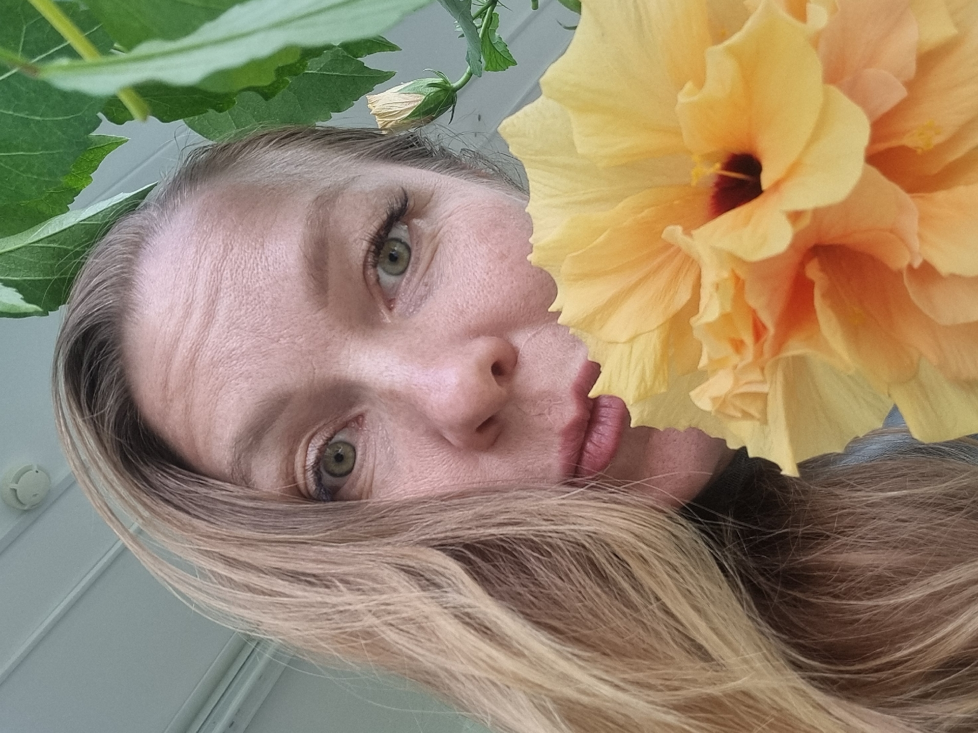 A low-angle, tilted, medium shot depicts a fair-skinned woman with blonde and brown highlighted hair, her face partially obscured by a large, yellow hibiscus flower. The woman has green eyes and reddish-pink lipstick. Part of her face is visible: her left eye and cheek, nose, and lips. Some green leaves are visible at the top of the frame, obscuring the top of her head. A smoke detector is visible in the background, to the left of the woman's head. The scene is likely indoors. The location is not verifiable as Kemul, Russia.