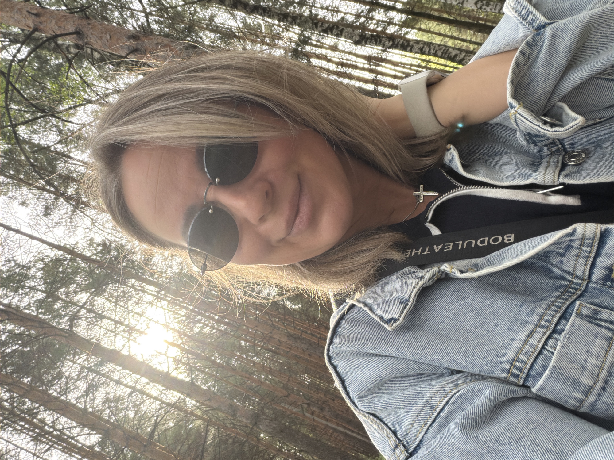 A selfie taken outdoors shows a woman with blonde hair wearing sunglasses and a denim jacket. The background suggests a forest with tall, slender trees, likely located in or around the city of Chaikovsky, Russia, given the context provided. The sun is visible through the trees, creating a lens flare effect. The woman is also wearing a smart watch. A silver necklace with a cross is visible on her neck, and a black strap with the inscription "BODULEATHE" is partially visible.