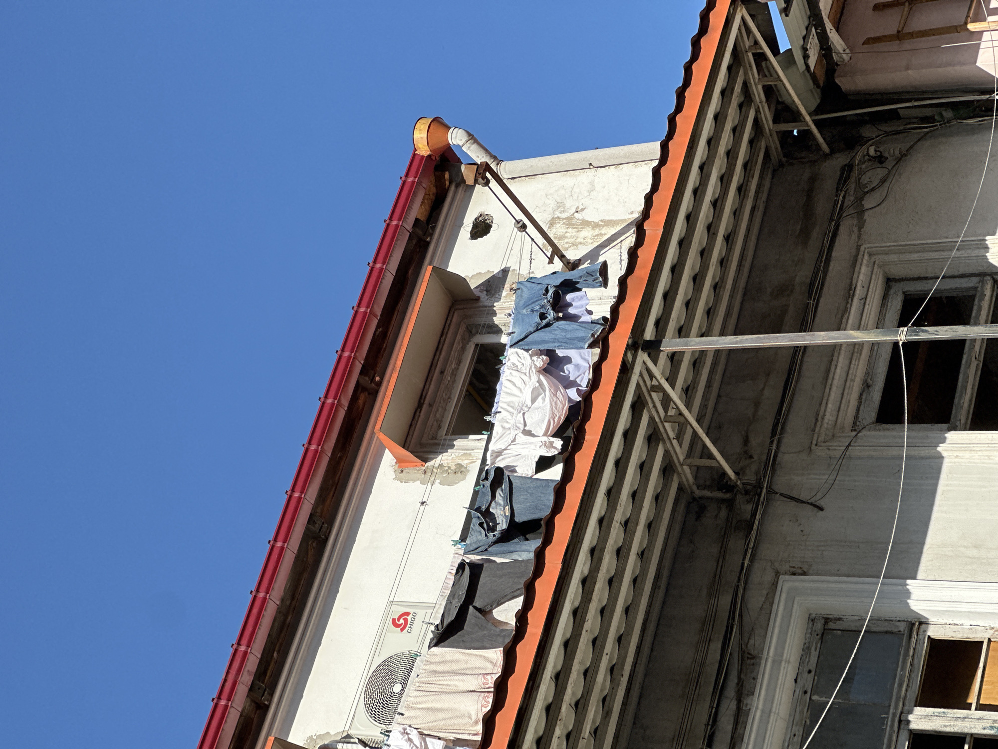 Low-angle view of an exterior facade of a white building, possibly in Batumi, Georgia, given the Chigo air conditioner visible. Clothes are hung out to dry along a makeshift line between the building's gutter and a structural support. The building has a red-tiled roof and ornate trim. The sky is clear and blue. Windows appear old and some are broken. Electrical wiring is visible along the building's exterior.