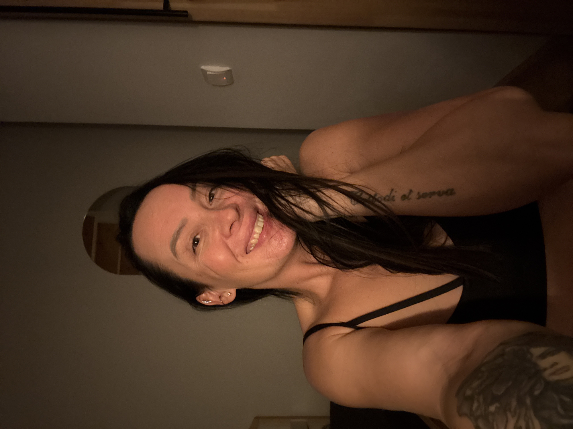 A woman with dark hair and tattoos on her arms is smiling at the camera. She is wearing a black top with thin straps. She appears to be indoors, possibly in a dimly lit room. A light fixture is visible on the ceiling. No information is available about the location in Chaikovsky, Russia.