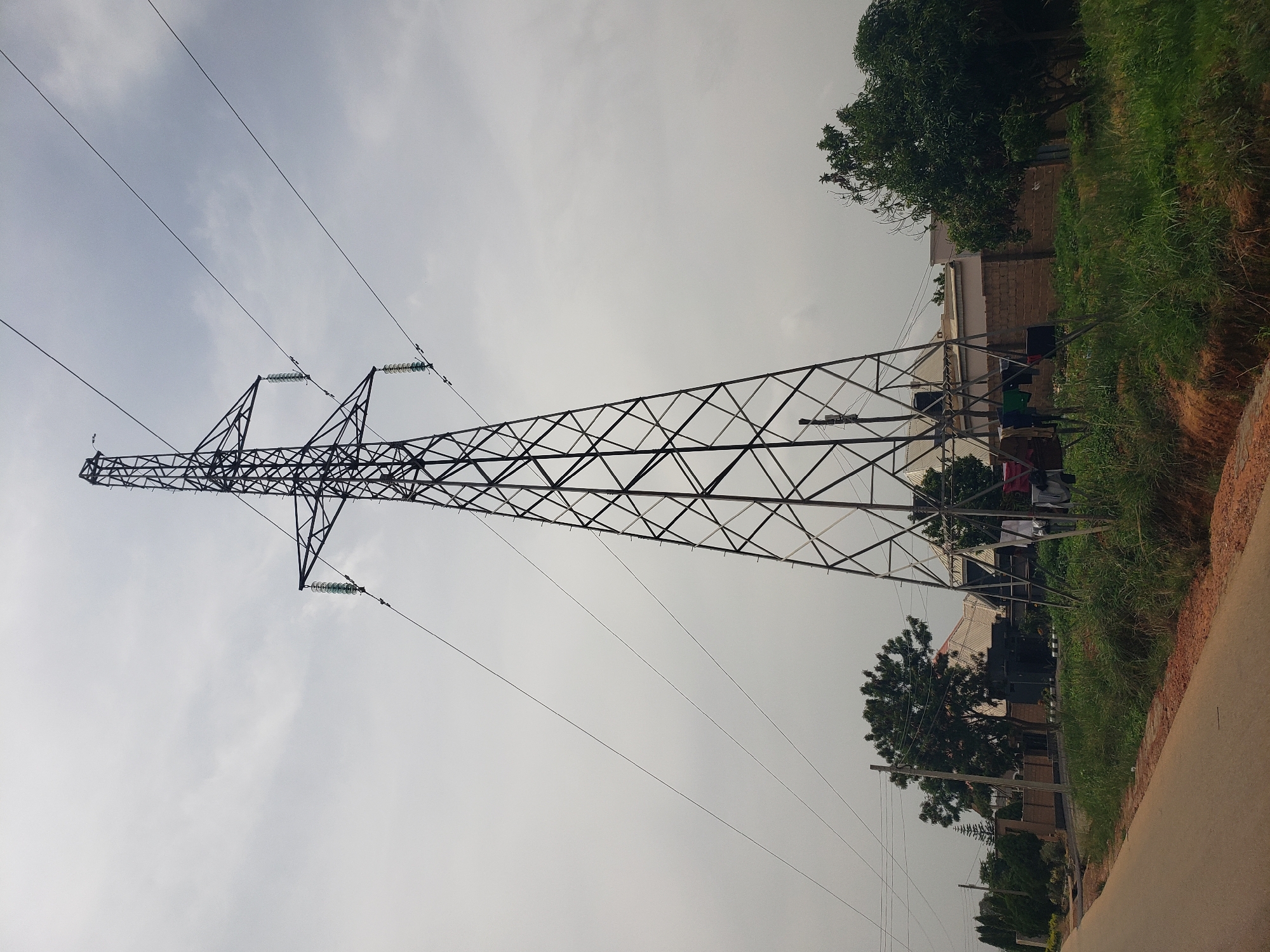 An exterior, low-angle, full shot captures a tall, black metal electricity pylon dominating the frame under an overcast sky. Three thick electrical cables extend from the pylon to the right, disappearing out of frame. Below the pylon, clothes are hung on a line, with several items visible. In the background, to the left of the pylon, a multi-story building with a light-colored roof and brick walls is partially visible, surrounded by trees. To the right of the pylon, more trees and a brick wall with a building are present. A paved road with a dirt shoulder runs along the bottom left of the image. The scene is likely located in a semi-urban or rural area within Rayfield, Nigeria, given the presence of residential structures, vegetation, and essential infrastructure like the electricity pylon.