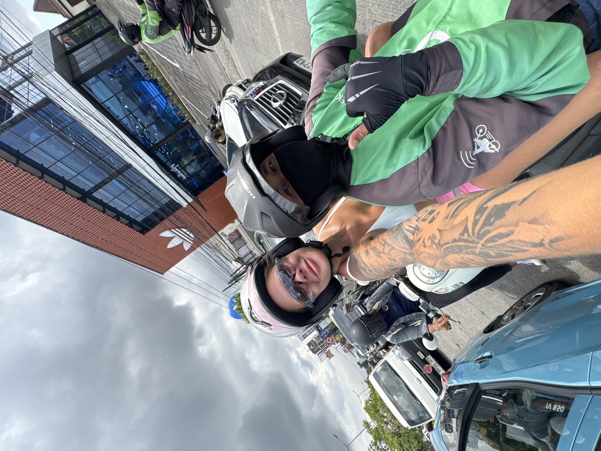 Two individuals are situated on motorcycles in what appears to be a bustling urban setting. One individual, wearing a pink helmet and sunglasses, is positioned in the foreground. The other individual, donned in a black helmet and a green jacket, is seated behind, appearing to be the motorcycle driver.
The location can be identified as Kuta, Indonesia, based on the presence of local landmarks. In the background, an Adidas store is visible. The surrounding environment includes buildings, cars, and other motorcycles, suggesting a busy street scene. The weather appears overcast, indicated by the cloudy sky overhead. Both individuals seem to be taking a selfie.