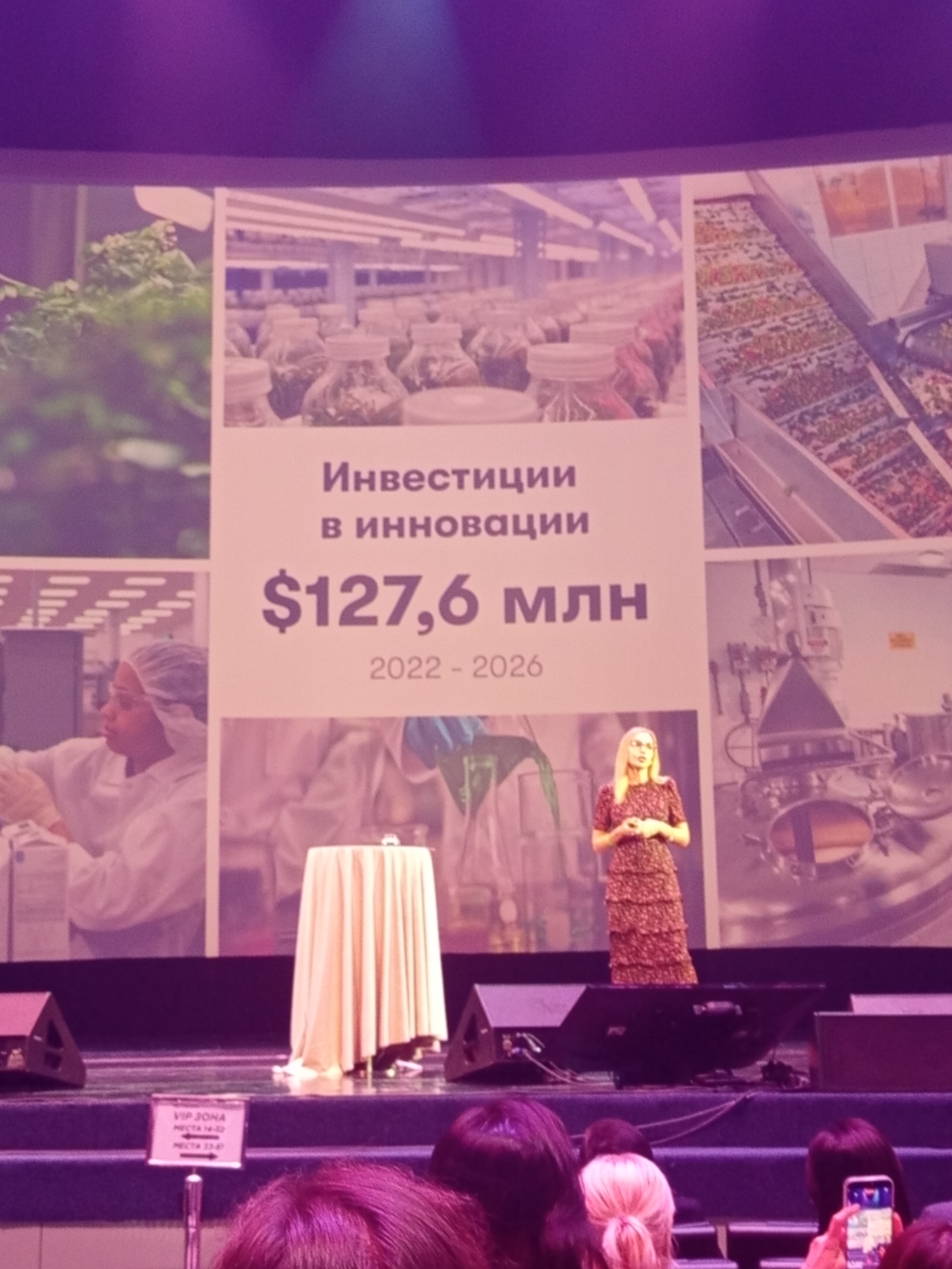 The image shows a large screen displaying a presentation with text in Russian. The text reads "Инвестиции в инновации" (Investments in innovation) and "$127,6 млн" ($127.6 million) along with "2022 - 2026". The screen is divided into several panels featuring images related to scientific research and development, including what appear to be laboratory settings with plants in jars, a conveyor belt with produce, and a woman in a lab coat. A woman in a patterned dress is standing on a stage in front of the screen, speaking to an audience. In the foreground, there are speakers and a sign that reads "VIP ЗОНА" (VIP ZONE). The overall setting suggests a conference or a business event. The location is not specified in the image.