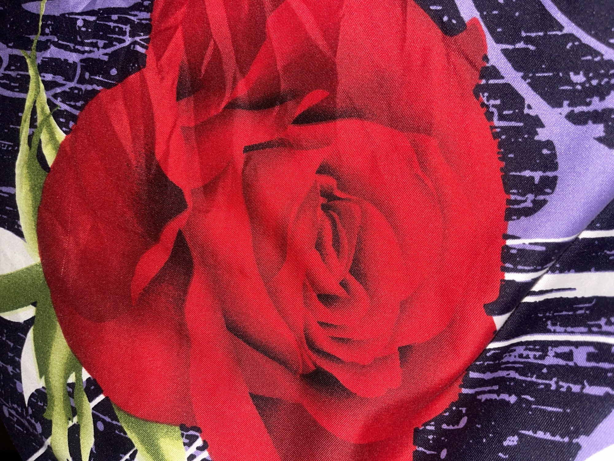 The image shows a close-up, top-down view of a fabric with a floral pattern. A large, stylized red rose dominates the center of the frame, with its petals appearing soft and rounded. Behind the rose, there is a background pattern featuring abstract shapes in black, purple, and white, interspersed with green leaves. The fabric appears to be silk or a similar material with a slight sheen. The image does not contain any information about the city of Minsk, Belarus.