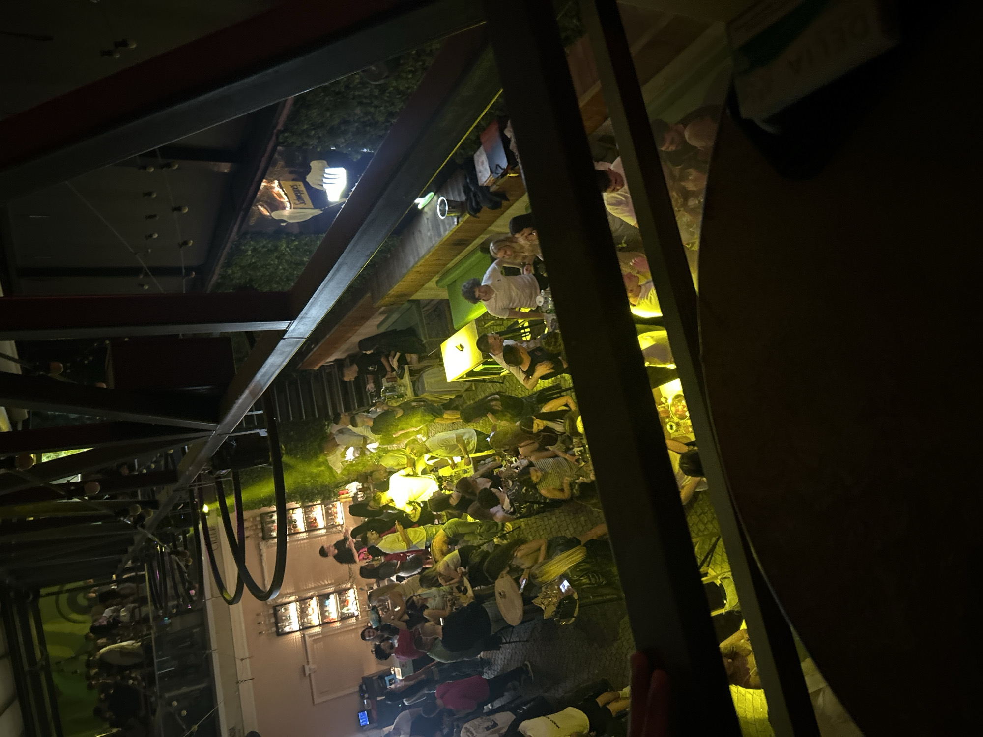The image shows a crowded indoor/outdoor venue, likely a bar or nightclub, with multiple levels and an open-air section. In the foreground, a dark wooden railing is visible, with a blurred table in front of it. Beyond the railing, the scene unfolds.
On the lower level, there is a dense gathering of people standing and sitting at tables, illuminated by bright yellow lighting. A bar area is visible with shelves of bottles. People are dressed casually in typical evening wear.
Above this, an elevated walkway or balcony overlooks the main gathering. More people are present on this level, some standing and observing, others interacting. A prominent structure of dark metal beams crisscrosses the upper part of the frame, suggesting industrial-chic architecture.
Further into the background, a sign for "Schweppes" is visible, illuminated with a neon glow and set against a backdrop of greenery, possibly a living wall. String lights are also visible in the distance, contributing to the ambient lighting. The overall impression is a lively social gathering in a modern, multi-level establishment.
The scene is consistent with venues found in the Kapana creative district of Plovdiv, Bulgaria, known for its vibrant nightlife, bars, and restaurants, often featuring multi-level spaces and industrial design elements.