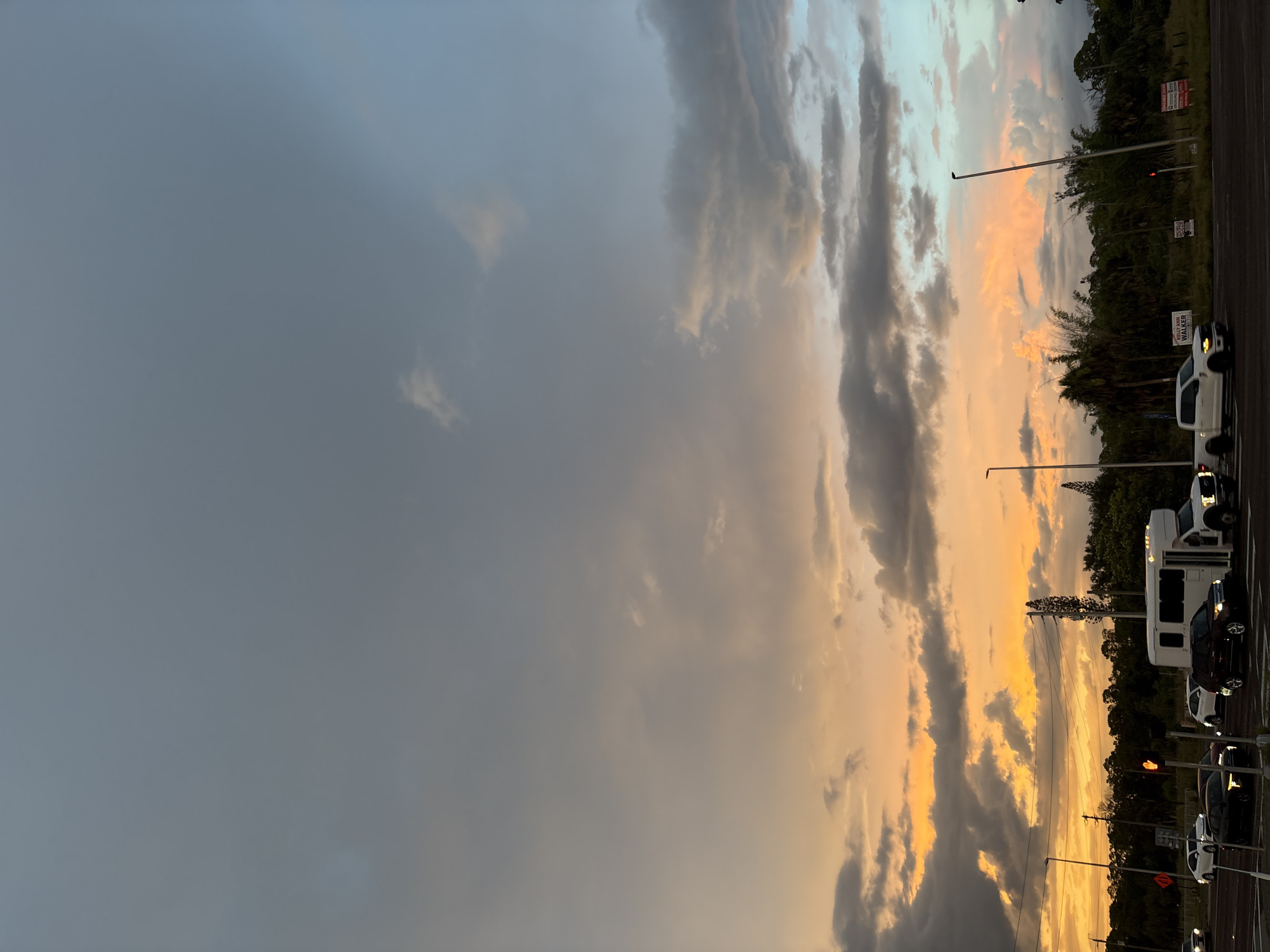 The image depicts a twilight scene on a road in Venice, Florida, United States, with a dramatic sky painted in hues of orange, pink, and grey. A traffic intersection is visible, with several vehicles including a white passenger van, a white pickup truck, and several sedans. The vehicles are stationary, indicating they are either waiting at a traffic light or stopped in traffic. The sky is filled with dynamic clouds, some illuminated by the setting sun with vibrant orange and pink tones, while others are dark and foreboding. A line of trees and vegetation forms the horizon behind the road, with some taller trees and utility poles interspersed. Several signs are visible along the roadside, though their text is not legible. The overall impression is of a natural phenomenon, the sunset, interacting with the man-made environment of a street in a semi-urban or suburban setting.