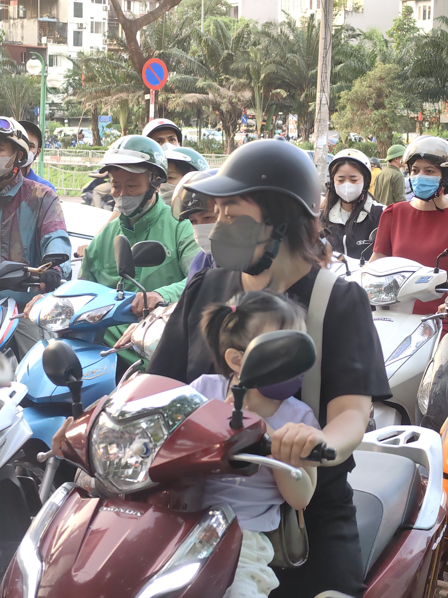 This image depicts a bustling street scene in Hà Nội, Vietnam, characterized by heavy motorbike traffic. The setting is an urban environment, likely during late afternoon or early evening given the soft, warm light.
In the foreground, a woman wearing a black short-sleeved top, a dark helmet, and a grey face mask is seen riding a red Honda scooter. A small child, possibly her daughter, sits in front of her, also wearing a purple face mask and a light-colored top with pigtails. Both appear to be navigating through the crowded street.
Behind them, a man in a distinctive green Grab uniform, wearing a matching green helmet with "Grab" partially visible, and a grey face mask, is positioned on a light blue Honda scooter. He, like many others, seems to be stopped or moving slowly in traffic.
Numerous other people populate the background, mostly riders or passengers on various scooters (white, blue, and other colors), almost all wearing helmets and face masks. One white scooter clearly displays the "Honda" brand name on its front. Further in the background, a man in an olive green military-style uniform is visible.
The scene is framed by typical urban elements: multi-story buildings in the distance, abundant palm trees and other foliage, and a utility pole. A prominent red circular traffic sign with a white horizontal bar, indicating "No Entry" or "Do Not Enter," stands on a pole in the upper left. The weather appears clear. The overall impression is one of a busy, everyday commute or movement within a vibrant Vietnamese city.