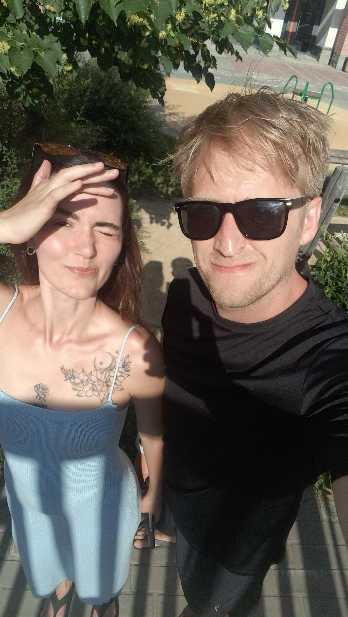 A man and a woman are taking a selfie outdoors, possibly in Tyumen, Russia, with trees and a building visible in the background. The woman, on the left, is wearing a light blue dress and has a tattoo on her chest. She is shielding her eyes from the sun with her hand. The man, on the right, is wearing a black t-shirt and sunglasses. They are both looking at the camera. The lighting suggests it is a sunny day.