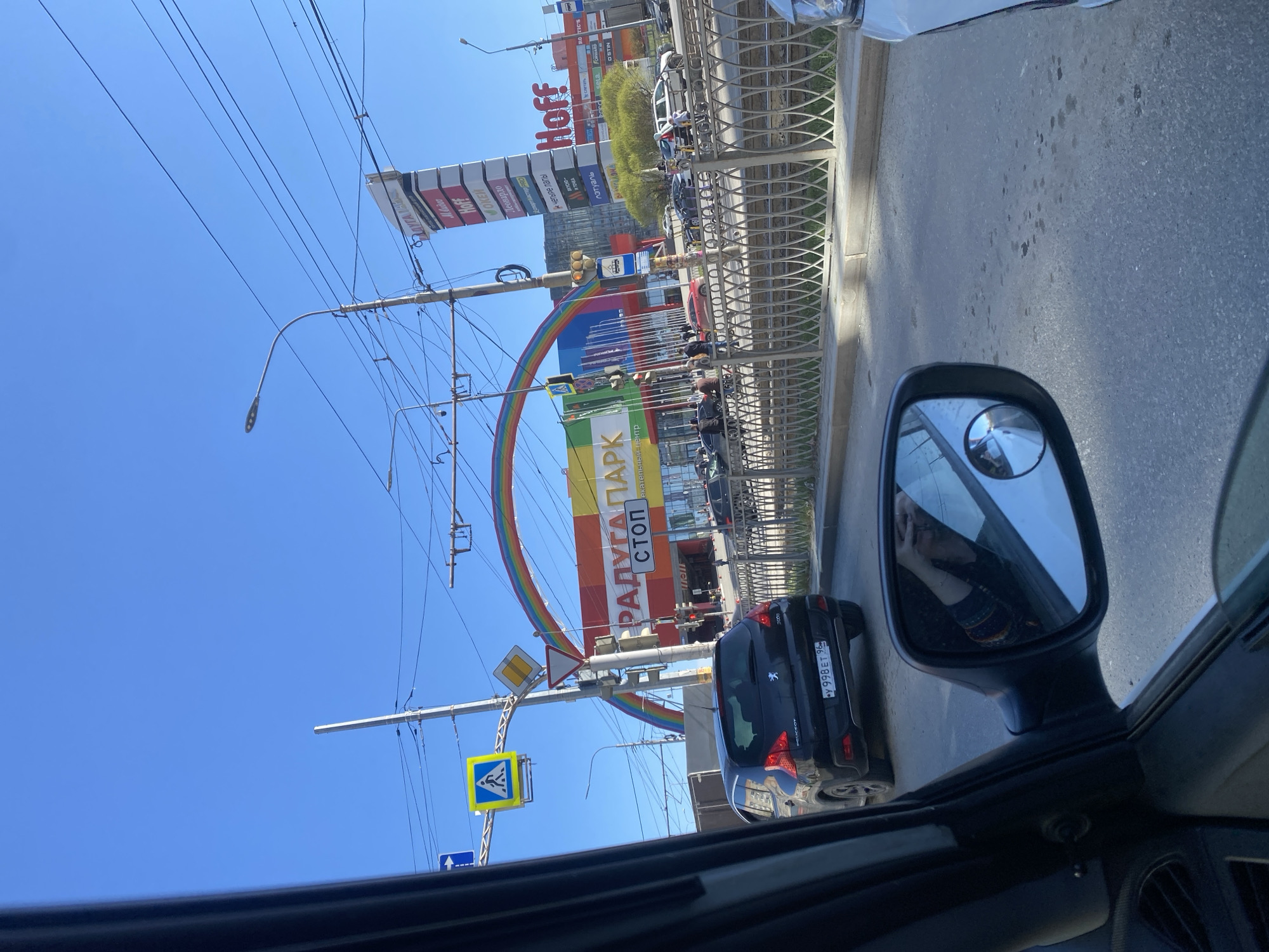 Here is a description of the image:
The image is taken from inside a car, likely stopped in traffic, looking out towards a busy street in Yekaterinburg, Russia. The rear-view mirror is visible, reflecting the driver. The focus of the shot is a large, colorful rainbow arch that is set up over the street. The rainbow spans across a street intersection, marking the entrance to the "Raduga Park" entertainment center. The arch is flanked by street signs and lamp posts.
Behind the rainbow is a tall building with multiple billboards including the "Hoff" brand. The license plate on the black Peugeot behind the car reads "Y 998 ET 96", a plate from the Sverdlovsk Oblast. The sky is clear and blue, suggesting a sunny day. Pedestrians can be seen crossing the street.