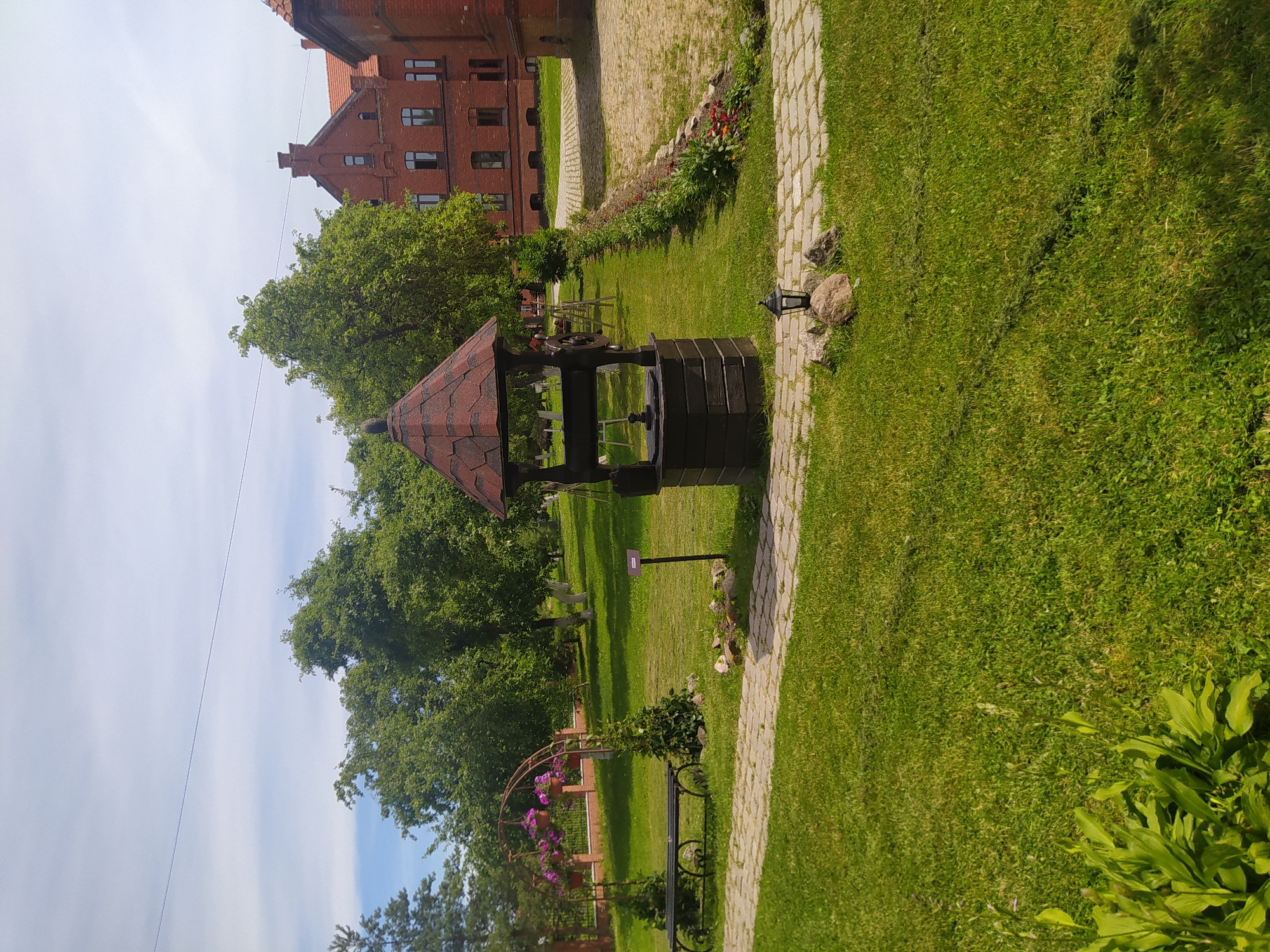 This is a full-shot, eye-level, outdoor photograph taken on a sunny day, featuring a well in the foreground. The well is made of dark wood and has a red, shingled roof. To the left of the well is a paved pathway made of stone. Beyond the pathway, a black metal bench is visible, with a decorative metal archway behind it, holding pink flowers in hanging baskets. A short, dark green bush is planted on either side of the bench. The background reveals lush green grass, trees with full foliage, and a large, red brick building with a tiled roof and multiple windows. The building appears to be historic, with a pitched roof and architectural details typical of older European structures. The sky is partly cloudy, with wisps of white clouds against a light blue background. The scene appears to be a well-maintained garden or courtyard area, possibly part of a historical site or estate. The location is identified as the Gusevsky Museum of Local Lore in Gusev, Russia.