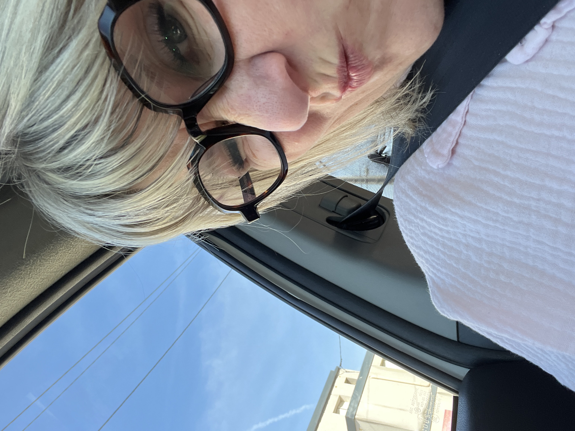 A woman with blonde hair and glasses is taking a selfie inside a vehicle. She is wearing a pink shirt and a black seatbelt is visible across her chest. The sky is visible through the car window, and a building can be seen in the distance. The photo was taken in Montélimar, France.