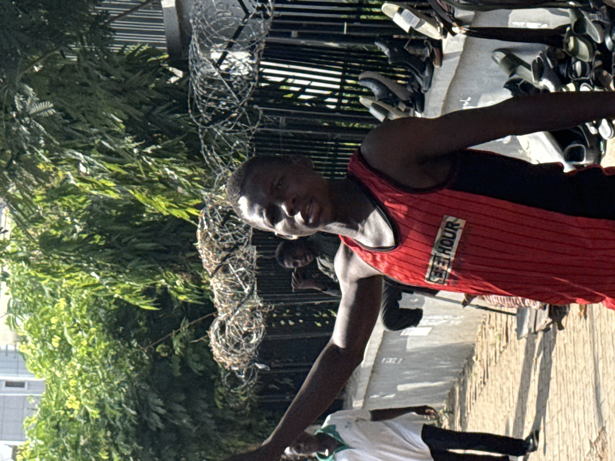 A medium shot captures a medium-skinned man wearing a red and black tank top. The foreground is composed of a barbed wire fence and green vegetation. In the background, a building is visible, with a glimpse of the sky. The setting appears to be an urban environment, possibly a street in Abuja, Nigeria. Several men are in the background.