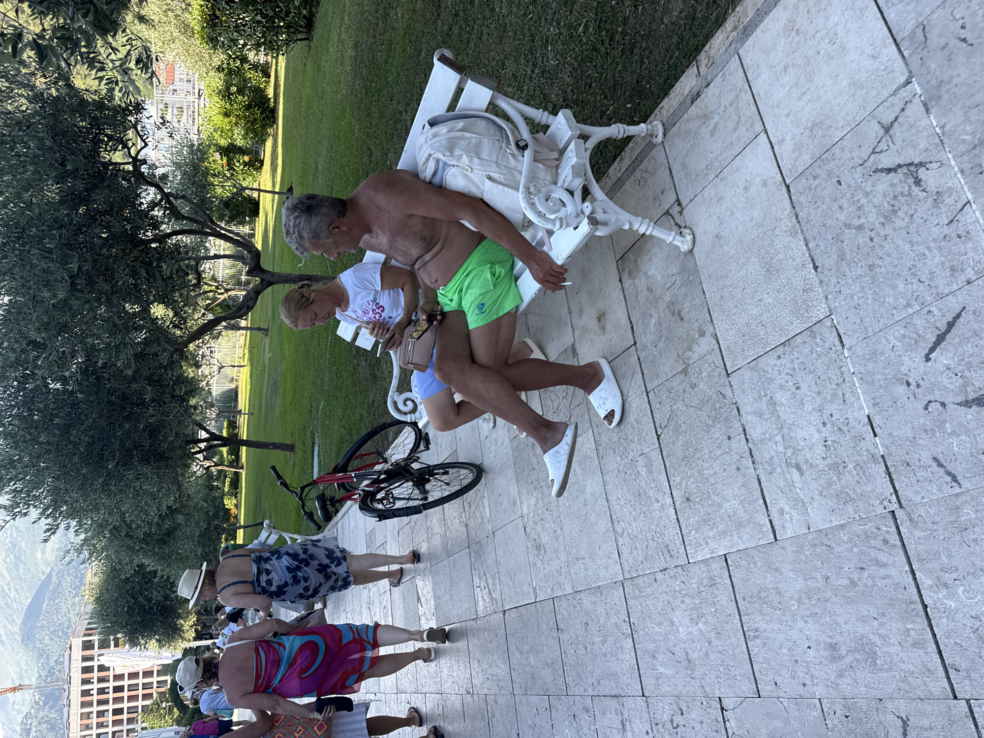 In Budva, Montenegro, a man and a woman sit on a white park bench in a grassy area adjacent to a paved walkway. The man, bare-chested and wearing green shorts, is holding a cigarette and looking to his right. Beside him on the bench is a light-colored backpack. The woman, wearing a white t-shirt and blue jeans, is looking down at her phone. To their left, a red bicycle is leaning against a metal fence. Behind them, several people are walking on the paved walkway, some wearing colorful sarongs and hats. In the background, olive trees and other vegetation are visible, with buildings and mountains further beyond. The paved area consists of large, light-colored stone tiles.