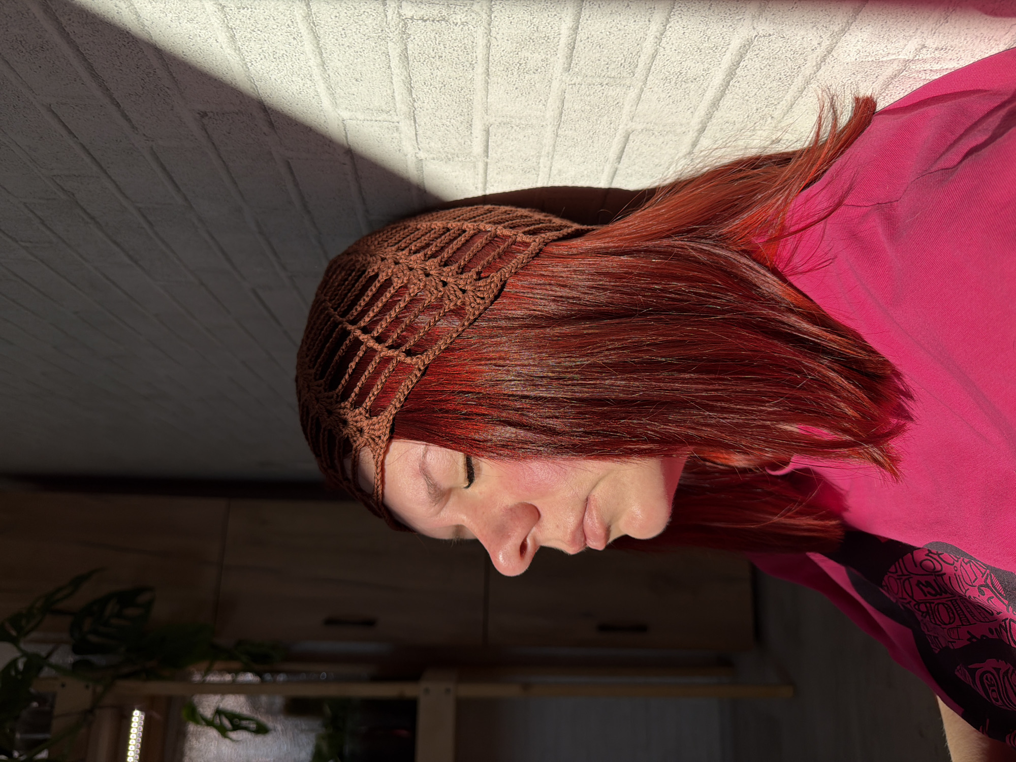 The image depicts a person with reddish-brown hair wearing a brown crocheted head covering. The person is wearing a bright pink shirt with black designs on the sleeve. They are turned to the side with their eyes closed, illuminated by sunlight. The background includes a white brick wall, partially obscured by a shadow, and a glimpse of wooden cabinets and green plants in a dimly lit interior. There is no indication to confirm it is Chaikovsky, Russia.