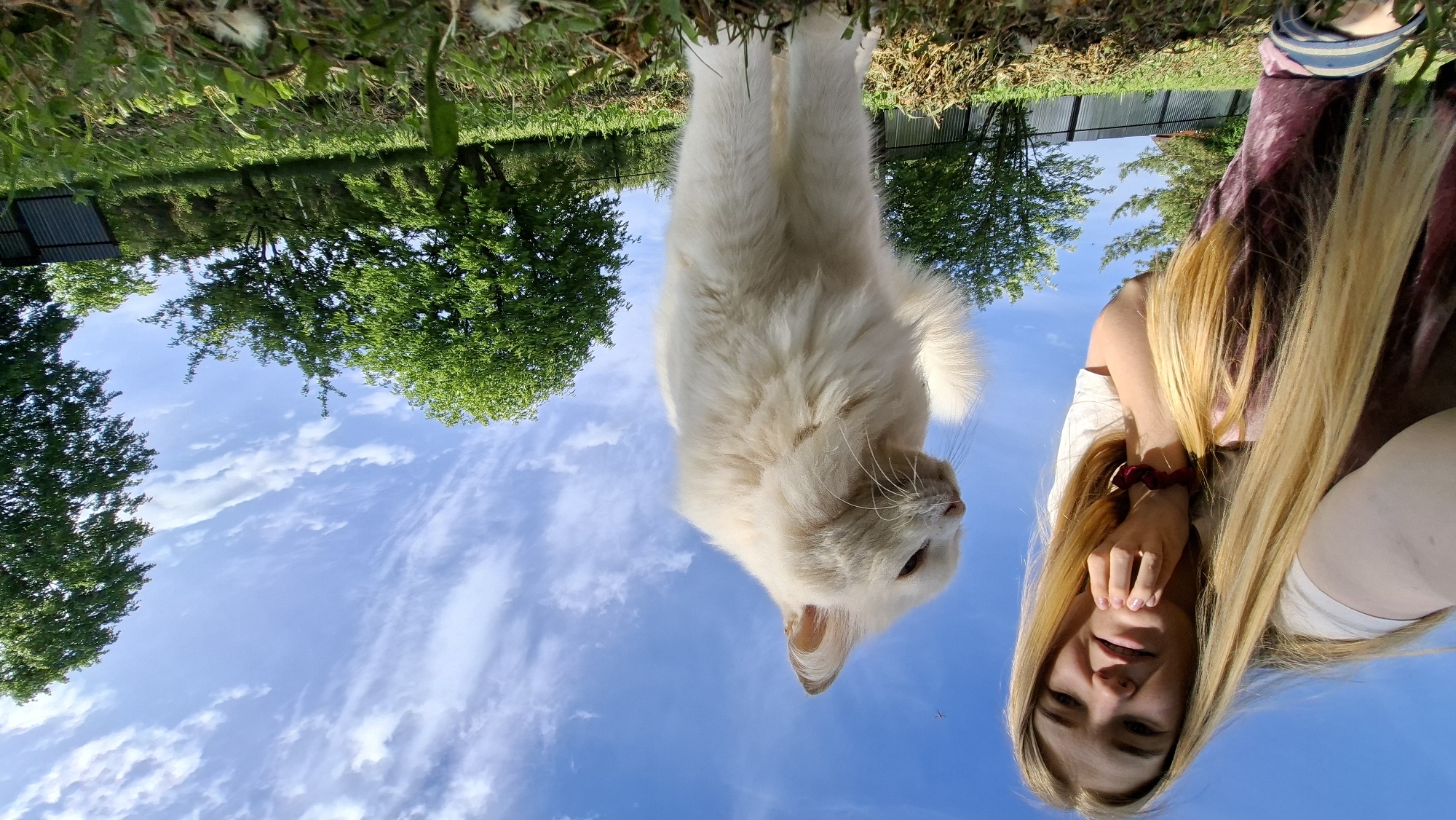 The image depicts a selfie taken in what appears to be a rural or semi-rural setting near Большой Букор, Russia. The sky is visible as a backdrop, partially obscured by tree foliage. In the foreground, a woman with long blonde hair is visible. She has her hand near her face. Beside her, a large white cat is prominent in the frame. The grass and more trees are also visible. The photo is taken at an unusual angle.