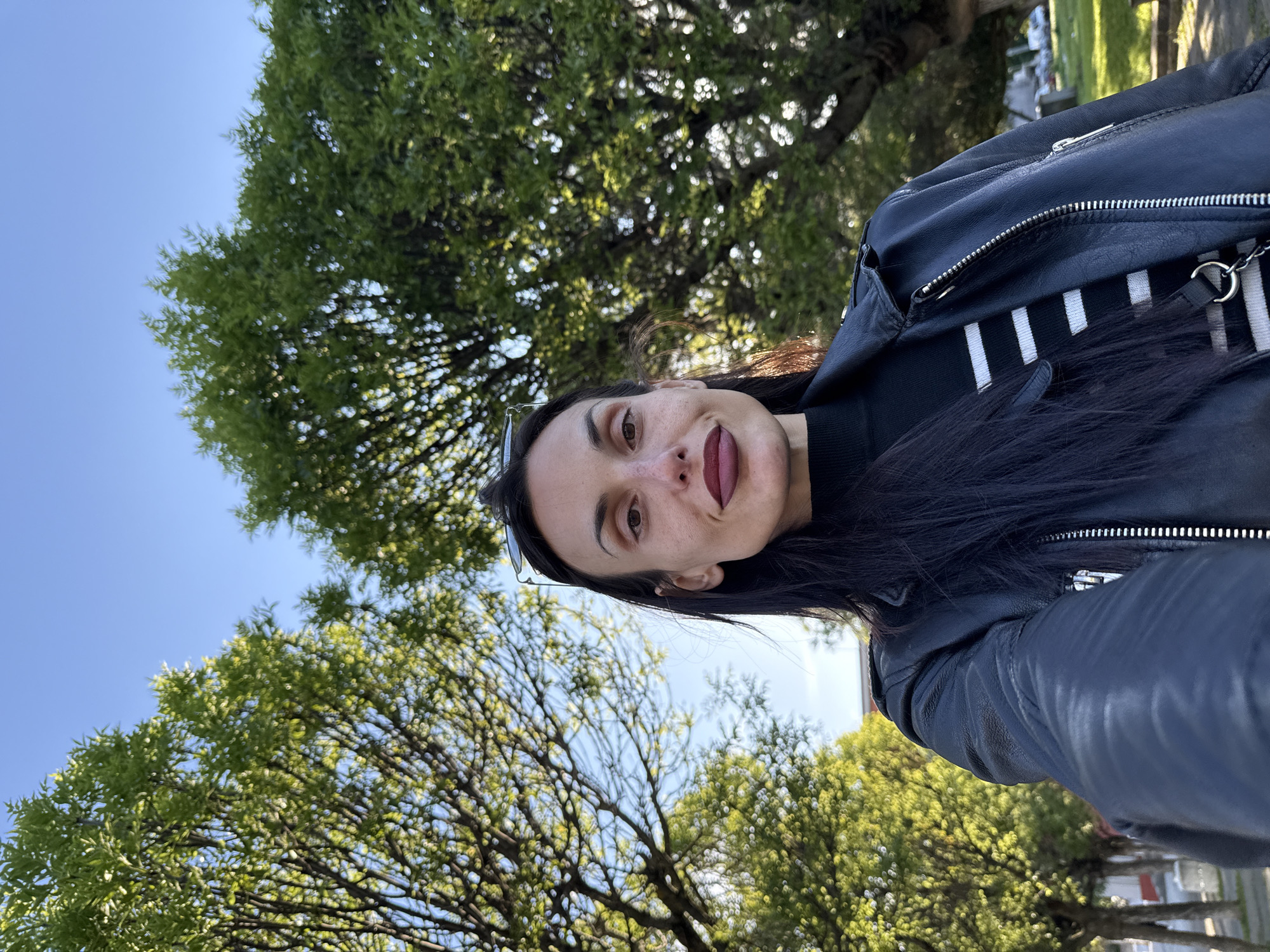 A woman is taking a selfie outdoors, likely in a park or green space. The photo is shot at an angle, with trees and blue sky visible in the background. The woman has dark hair and is wearing a black leather jacket over a striped shirt. She is sticking her tongue out slightly. The location is Pleven, Bulgaria, although specific landmarks are not visible.