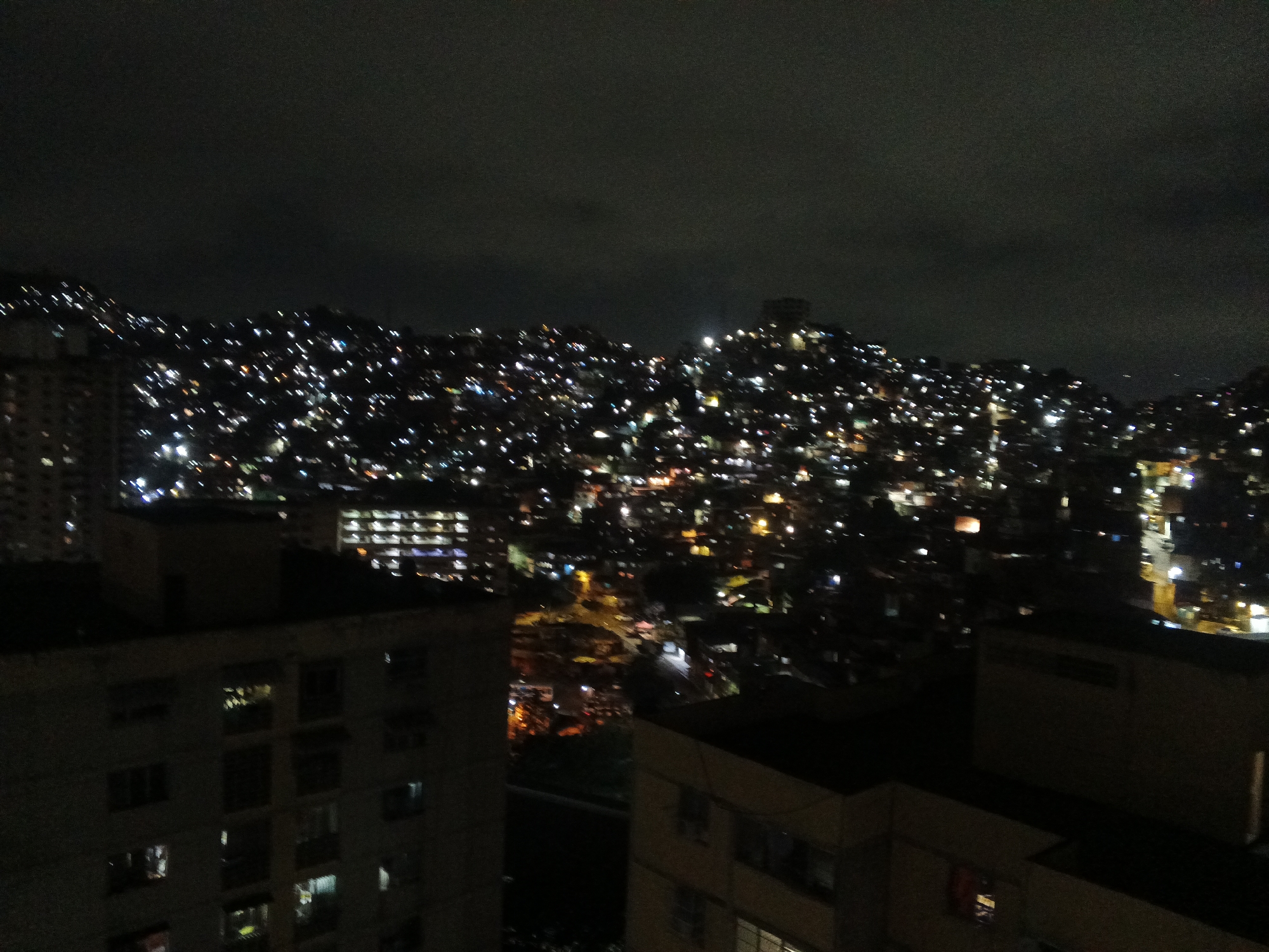 The image shows a night view of Caracas, Venezuela. The foreground features dark buildings with some lit windows. In the background, a densely populated hillside is visible, dotted with numerous lights that appear to be homes. The sky is dark and cloudy. The scene depicts the urban landscape of Caracas at night.