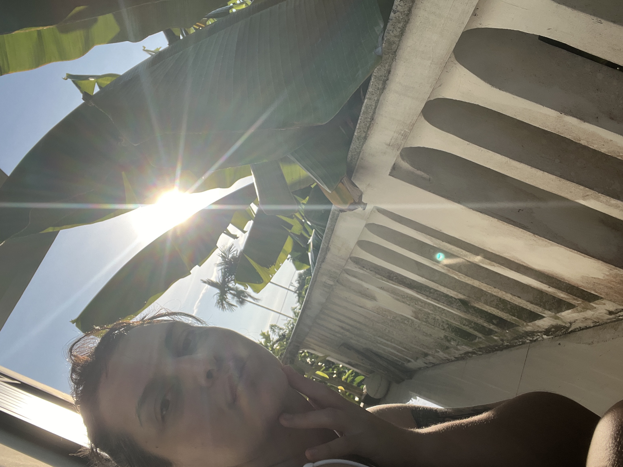 A first-person selfie is taken outdoors with the sun directly behind large green leaves, creating a lens flare effect. The person, whose face is partially visible in the foreground, is looking towards the camera and touching their chin. Behind the person and the leaves, a white balustrade with arched openings runs horizontally. In the background, palm trees and other foliage suggest a tropical or subtropical environment, consistent with Đà Nẵng, Vietnam. The sky is blue with some wispy clouds. The overall lighting is bright and sunny.