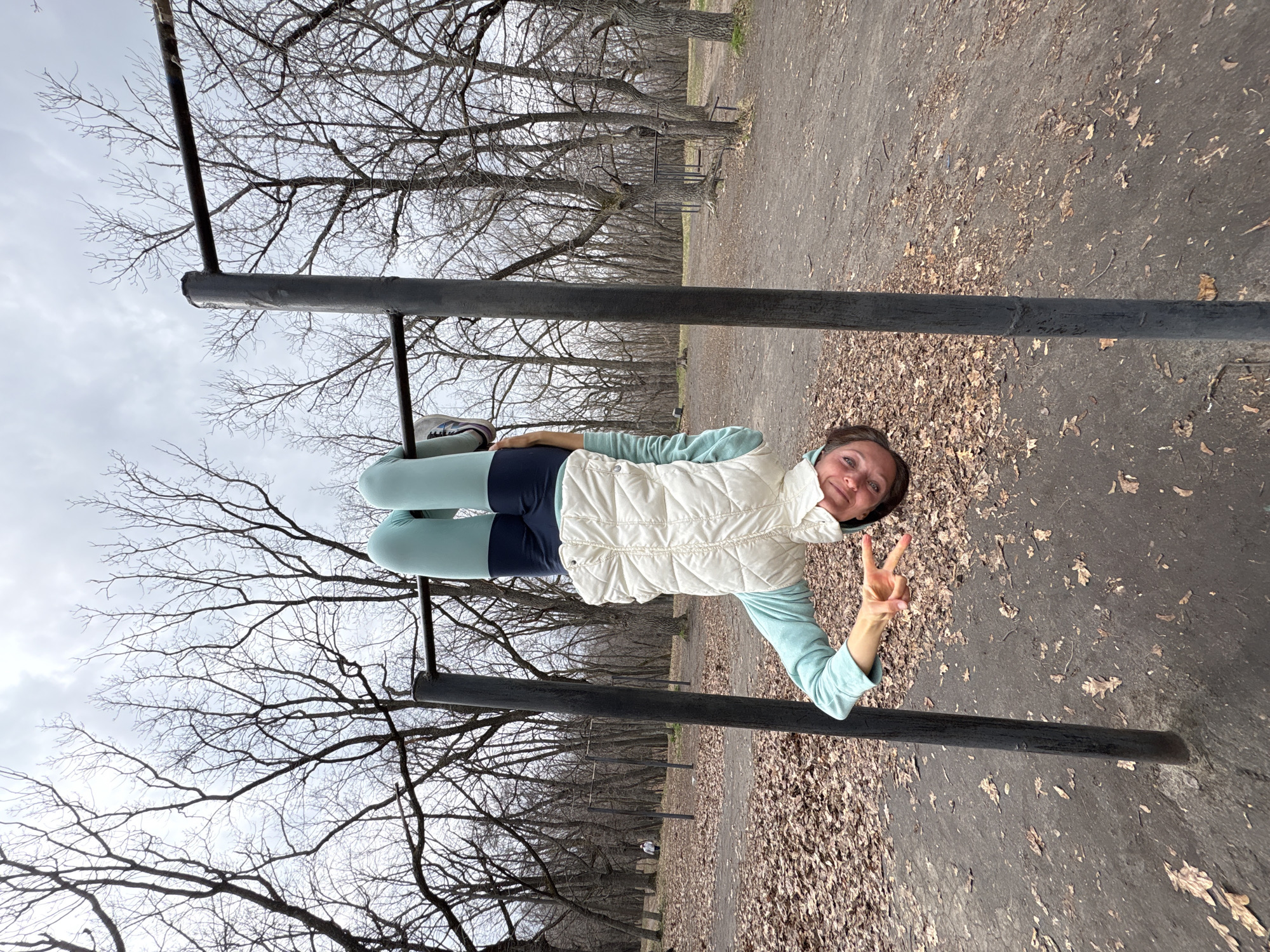 The image shows a woman standing between parallel bars in a park. She is wearing a light blue long-sleeved shirt, a white vest, dark blue athletic shorts, and light blue leggings. She is smiling and making a peace sign with her right hand. The park has dirt paths covered in fallen leaves. Bare trees are visible in the background. The location is likely a public park with exercise equipment. No specific information is available to confirm that this park is located in the city of Ulyanovsk, Russia.