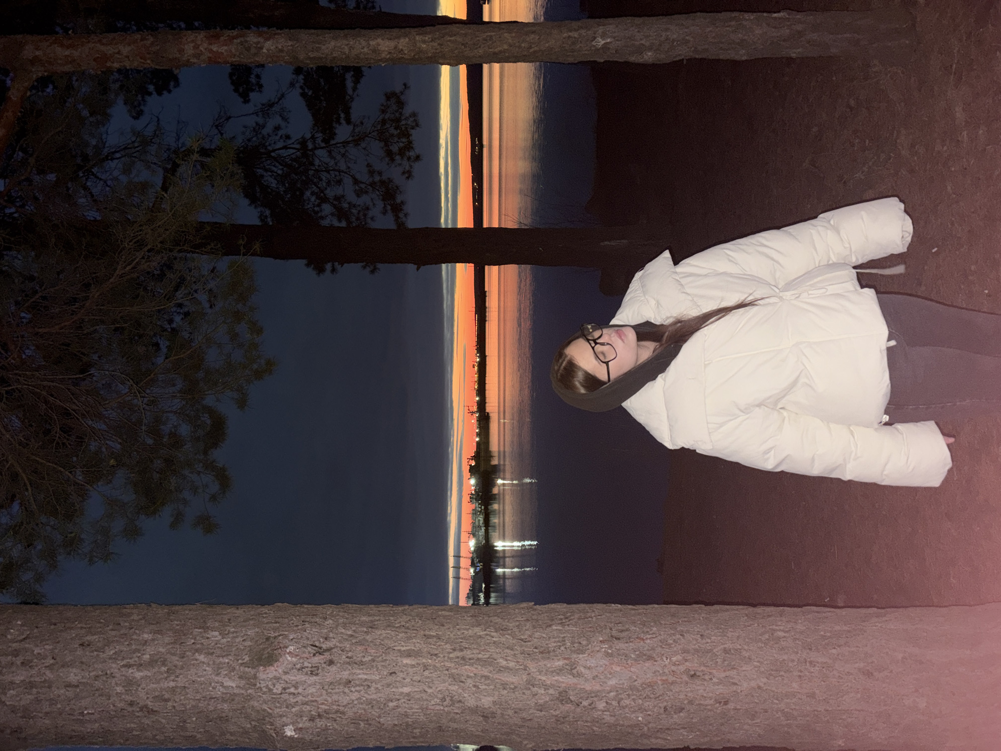 The image captures a tranquil twilight scene by a large body of water, possibly in the vicinity of Chaikovsky, Russia. The composition is framed by dark, silhouetted tree trunks and branches on the left and right sides.
In the mid-ground, a young woman is visible, wearing a light-colored, possibly white or cream, puffer jacket and eyeglasses. Her long dark hair flows down her back, and she appears to be looking towards the horizon, observing the view.
The notable feature of the scene is the sky at dusk, which presents a striking gradient of colors. Along the horizon, where the sun has recently set or is in its final descent, warm hues of orange, pink, and yellow dominate, gradually transitioning to deeper blues and grays higher in the sky. These vibrant colors are beautifully reflected in the calm waters, creating a mirror-like effect. Across the expanse of water, numerous small lights from a distant settlement or city are visible, twinkling against the deepening twilight. The overall atmosphere is one of peaceful contemplation. There is no visible text in the image.