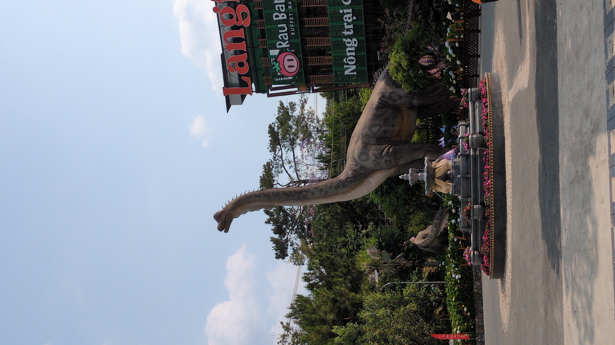 The image shows a life-sized dinosaur statue in front of a restaurant with a sign that reads "Làng Rau Ba Buffet, Nông trại C". The sky is partly cloudy. The scene is located in Cam Ly - Đà Lạt, Vietnam. A fountain is present next to the dinosaur. A road runs along the right side of the scene. Trees and other greenery are visible in the background.