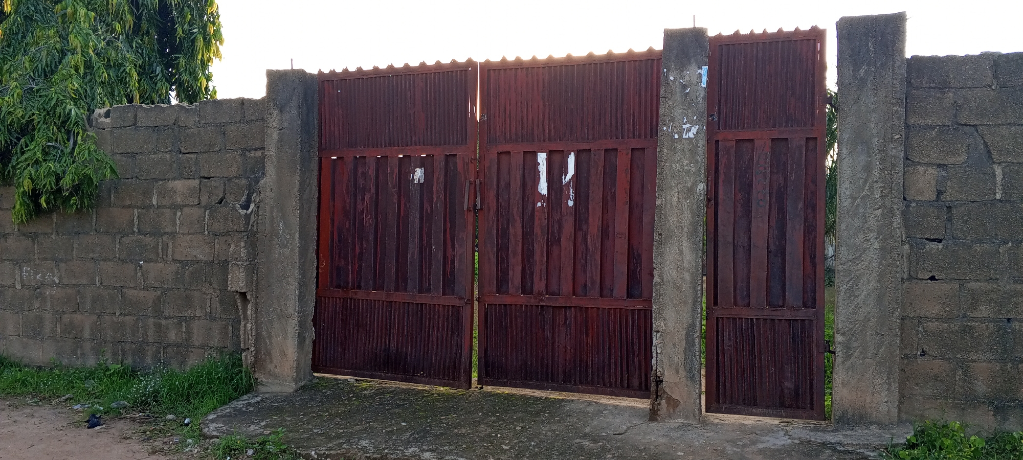 The image shows a weathered, dark red metal gate with vertical corrugated panels, set within a gray cinder block wall. The gate is divided into two main sections, with a smaller third section on the far right. The gate is supported by concrete pillars. To the left of the gate, the cinder block wall continues, with dense green foliage extending over the top. The ground in front of the gate is a mix of dirt and patches of concrete with some grass growing around the edges. The scene appears to be outdoors, possibly in a residential or industrial area, with the sky visible in the upper portion of the image. The overall impression is one of a functional, utilitarian entrance that has experienced some age and exposure to the elements.