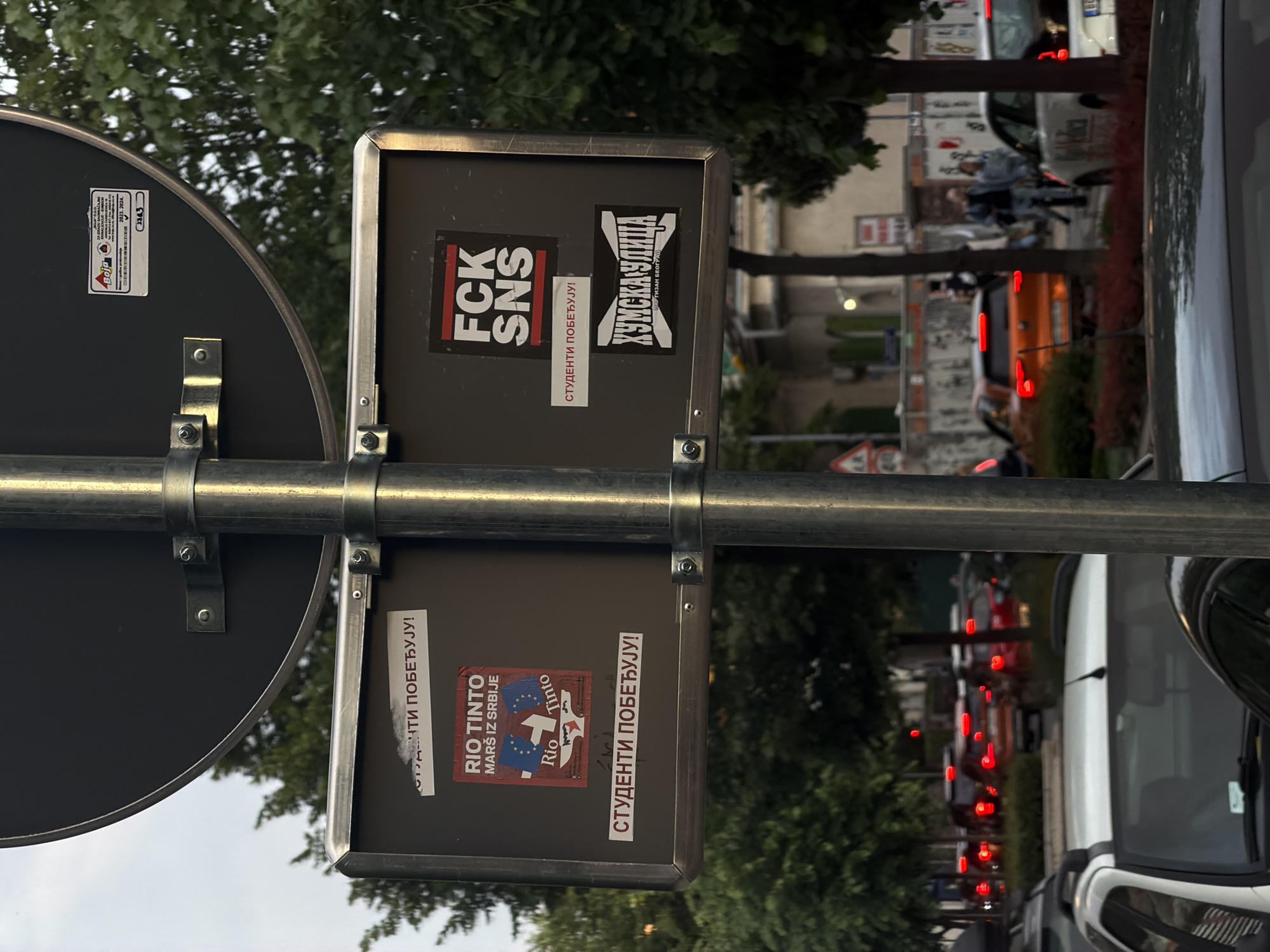The image shows a street sign in Vracar Urban Municipality, Serbia, covered with political stickers. The stickers feature anti-government slogans and messages related to the Rio Tinto mining project. The sign is attached to a pole with metal brackets. In the background, there is street traffic.