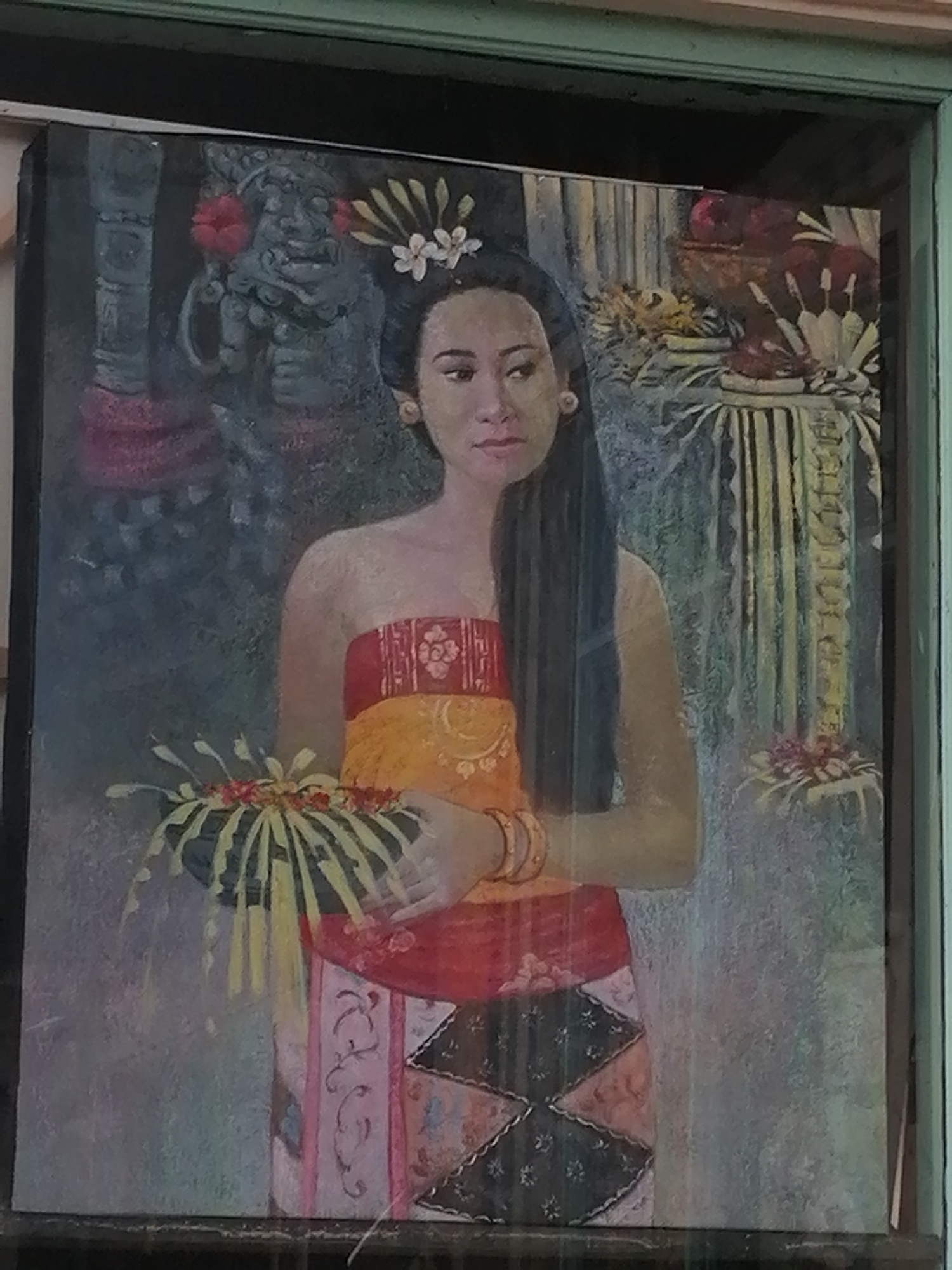 A traditional Balinese painting depicts a young woman adorned in ceremonial attire, holding a woven basket filled with offerings. Her dark hair is styled with flowers, and she wears ornate jewelry. The backdrop features elements of Balinese art and architecture, including a stone carving and floral arrangements, suggesting a temple or cultural setting. The painting is framed and appears to be displayed in a location within the Ubud District, Indonesia, a region renowned for its artistic heritage.