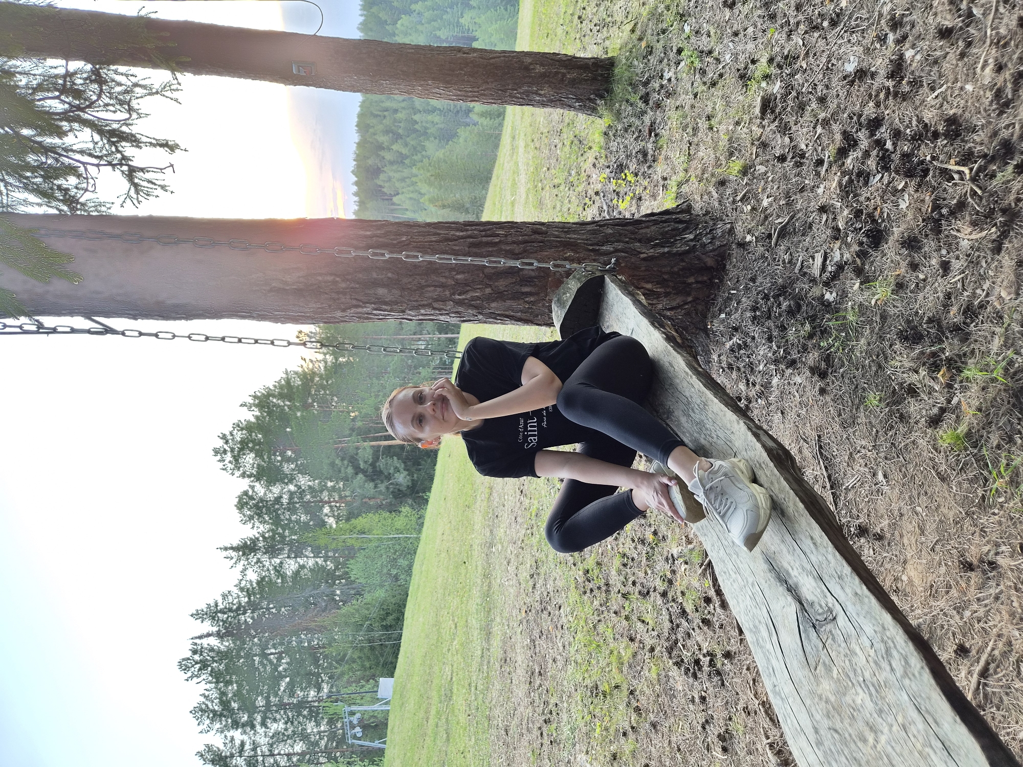 A photograph shows a young woman sitting on a wooden swing bench suspended by chains between two large pine trees in what appears to be a park or natural setting. The location is Прикамский, Russia.
The woman has fair skin and is wearing a black t-shirt and black leggings. She is sitting with her legs bent and her feet slightly elevated, with her hands near her face. She is wearing light-colored sneakers.
The wooden bench is aged and weathered, showing signs of wear. The area around the trees is covered in pine needles, small twigs, and other natural debris. Behind the trees, there is a view of a green forest and a sky that suggests either sunrise or sunset, with soft light filtering through the trees. The general impression is one of tranquility and a natural setting.