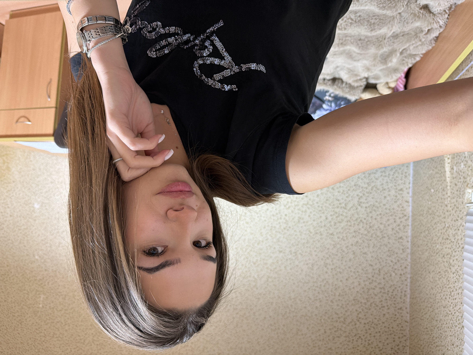 A photograph features a fair-skinned woman with light brown hair and blonde highlights, wearing a black t-shirt with sequin detailing. The image is oriented upside-down. She is wearing a watch and several bracelets on her left wrist and a ring on her finger. The background includes a textured wall and a cabinet with drawers. The exact location within the city of Sofia, Bulgaria, cannot be determined from the visual information provided.