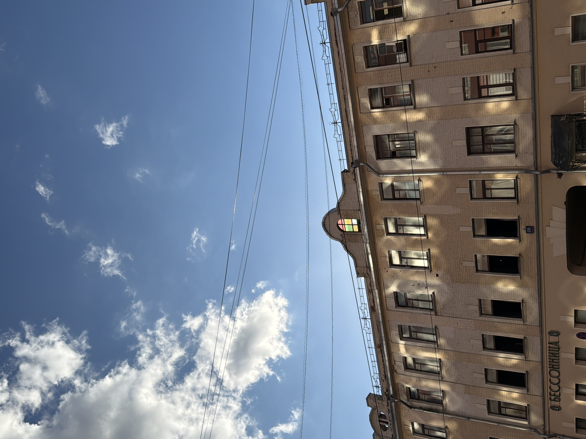 Here is a forensic description of the image:
The image is a low-angle shot of the exterior of a tall building, set against a sky with scattered clouds. The building's facade is light-colored, featuring a grid of windows that are mostly rectangular. The windows have dark frames, with some showing hints of reflections. There are power lines that run diagonally across the sky from upper left to the middle right of the image.
The sky is blue, populated by small puffy clouds at the bottom left and white, larger clouds at the middle left. The building appears to be located in a city, with its architectural style suggesting it's possibly in a European urban environment. There is text in an Cyrillic alphabet on the right side of the building. The building has a unique architectural feature at the top, including stained glass.